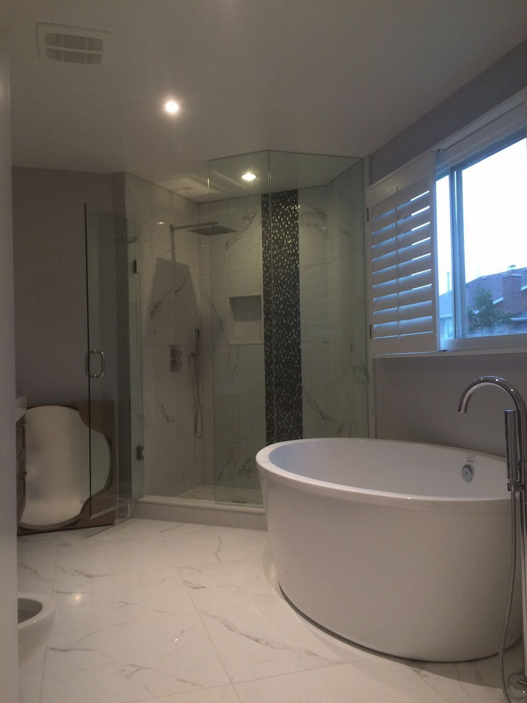 This Toronto bathroom renovation showcases a luxury layout featuring a freestanding bathtub, frameless glass shower enclosure, and marble-style flooring. The spacious design, modern fixtures, and natural light create a bright spa-like atmosphere. Upgraded lighting, premium finishes, and a functional layout enhance both comfort and daily usability, delivering a refined and contemporary bathroom space for modern living.