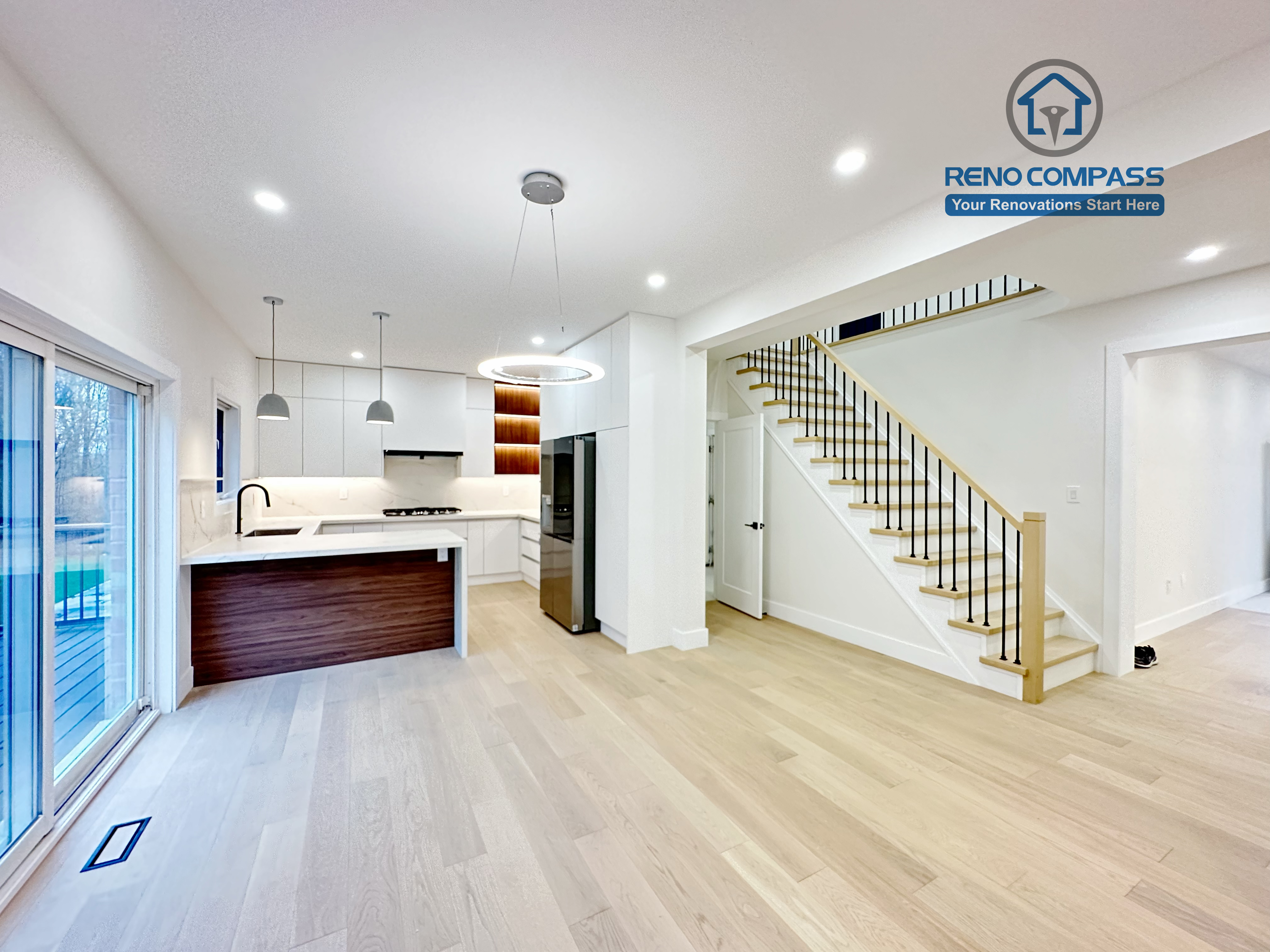 This image presents a modern kitchen renovation in Richmond Hill featuring full-height custom white cabinetry, a quartz waterfall island with wood panel detailing, and integrated stainless steel appliances. The design includes a built-in gas cooktop, minimalist pendant lights, recessed ceiling lighting, and light hardwood flooring for a clean, contemporary look. Large windows and patio access enhance natural light and openness. Completed by a professional Toronto Renovation Company, this kitchen renovation reflects refined craftsmanship, efficient layout planning, and modern family-focused design.