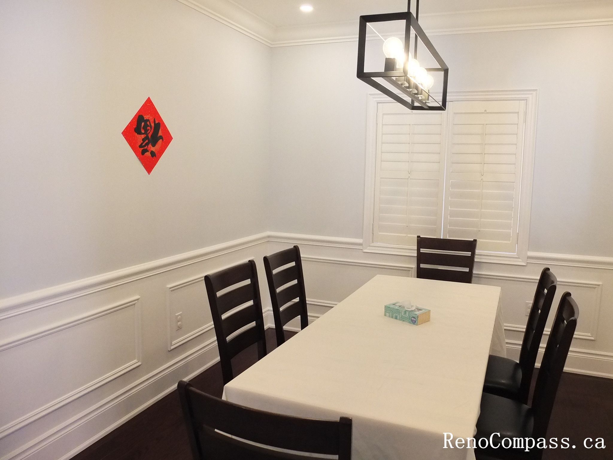 This Newmarket interior renovation features a clean and elegant dining room designed with classic wall wainscoting and rich dark hardwood flooring. Plantation shutters allow natural light to brighten the space while maintaining privacy, and the modern rectangular pendant light provides balanced illumination above the dining table. The simple layout and refined finishes create a comfortable setting for family meals and gatherings. Completed by Reno Compass Inc., this project highlights timeless interior design and high-quality craftsmanship for modern homes.