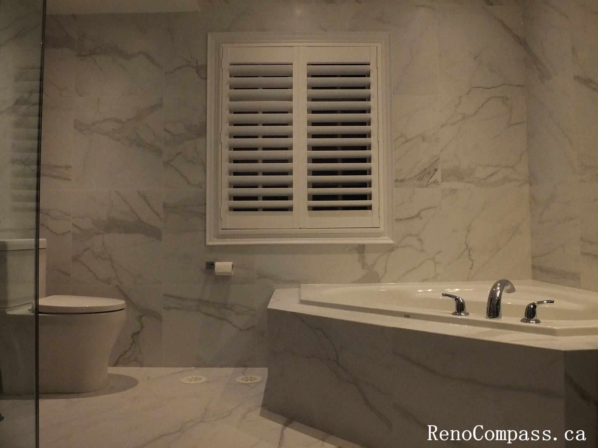 Modern luxury bathroom renovation in Unionville featuring marble wall tiles, a built-in soaking tub, and white plantation shutters.