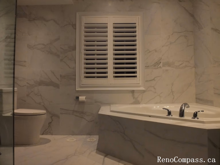 Modern luxury bathroom renovation in Unionville featuring marble wall tiles, a built-in soaking tub, and white plantation shutters.