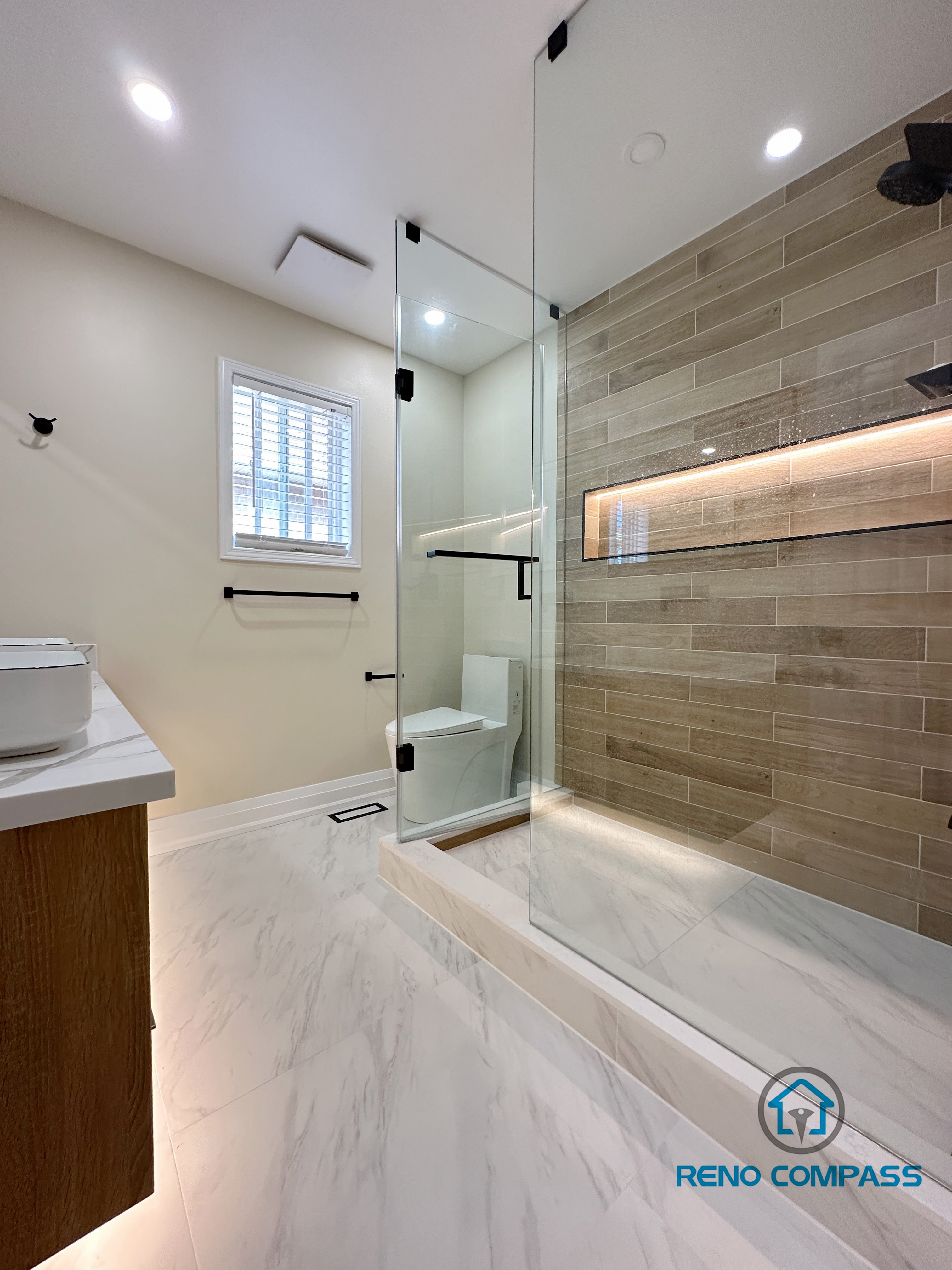 Contemporary bathroom renovation in Vaughan featuring a spacious walk-in shower with a frameless glass enclosure, custom LED-lit shower niche, and wood-look porcelain wall tiles. The shower design includes matte black fixtures, a low-profile shower base, and marble-look porcelain flooring for a seamless and modern finish. This project demonstrates high-quality workmanship and thoughtful layout planning by a trusted Toronto renovation company specializing in custom bathroom renovations.