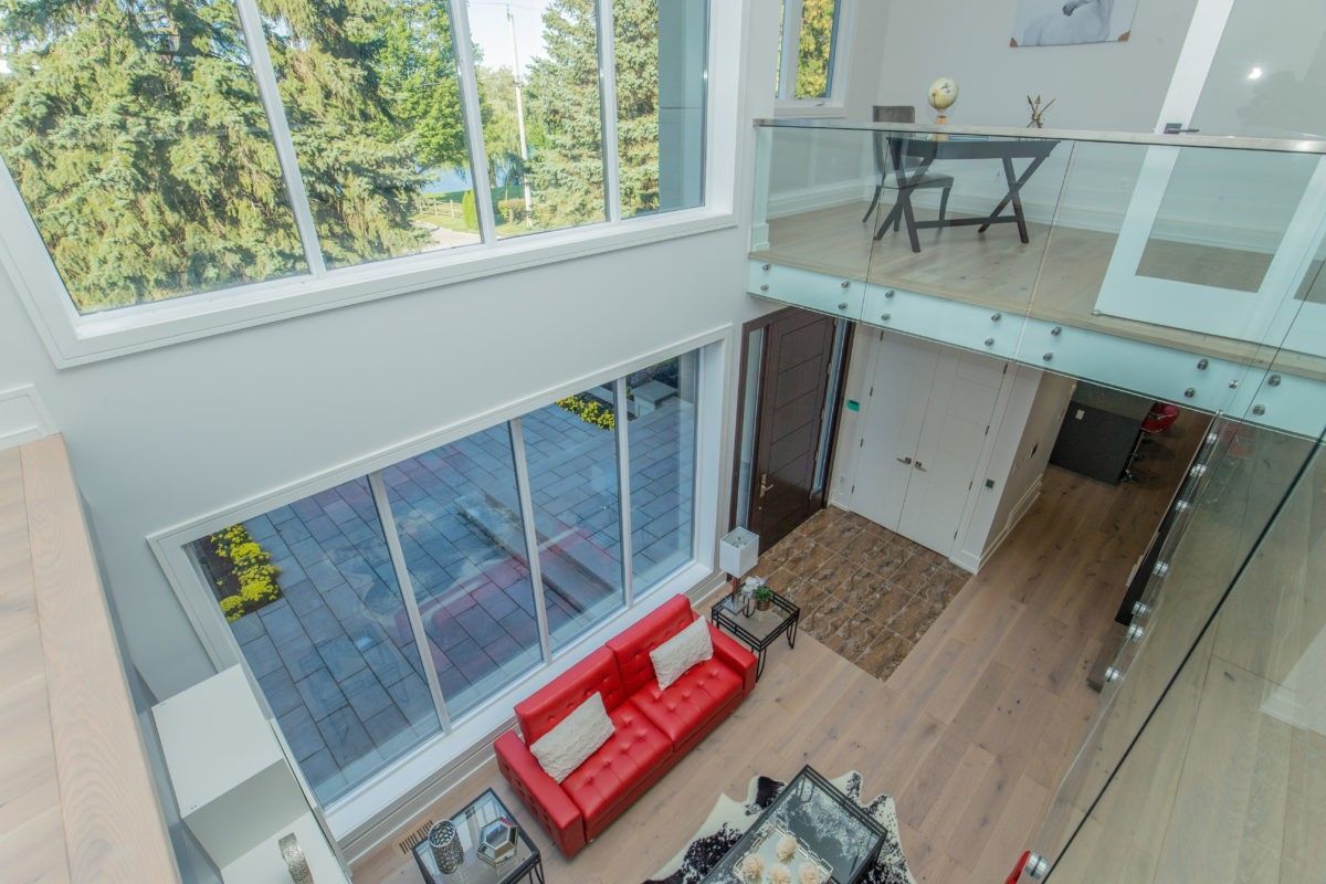Modern open-concept living room in a 5000 sqft Toronto home renovation featuring double-height ceilings, glass railing mezzanine, wide plank hardwood flooring, contemporary fireplace wall with built-in TV, and large sliding doors leading to a backyard deck. The space combines natural light, clean architectural lines, and bold red leather sofas to create a bright and sophisticated interior. Renovated by Reno Compass Inc., a professional Toronto renovation company specializing in large-scale custom home interior transformations.
