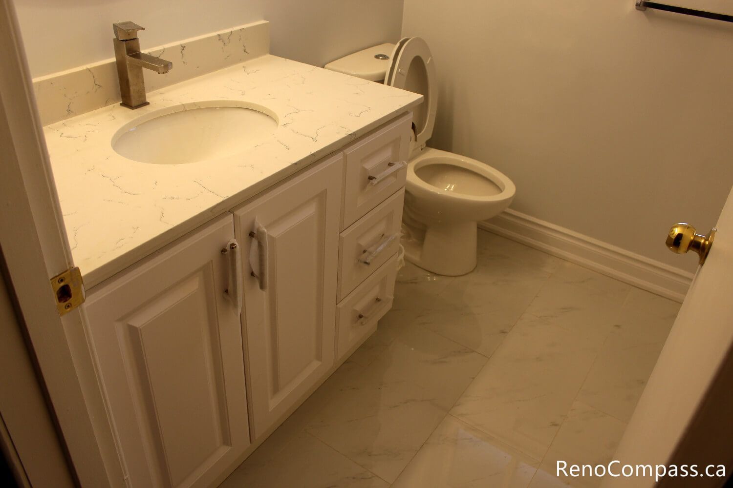 A bright bathroom renovation in Oshawa featuring a classic white shaker-style vanity with a quartz countertop, brushed gold faucet, and polished marble floor tiles. Expert bathroom remodeling by RenoCompass.ca to maximize small space functionality and style.