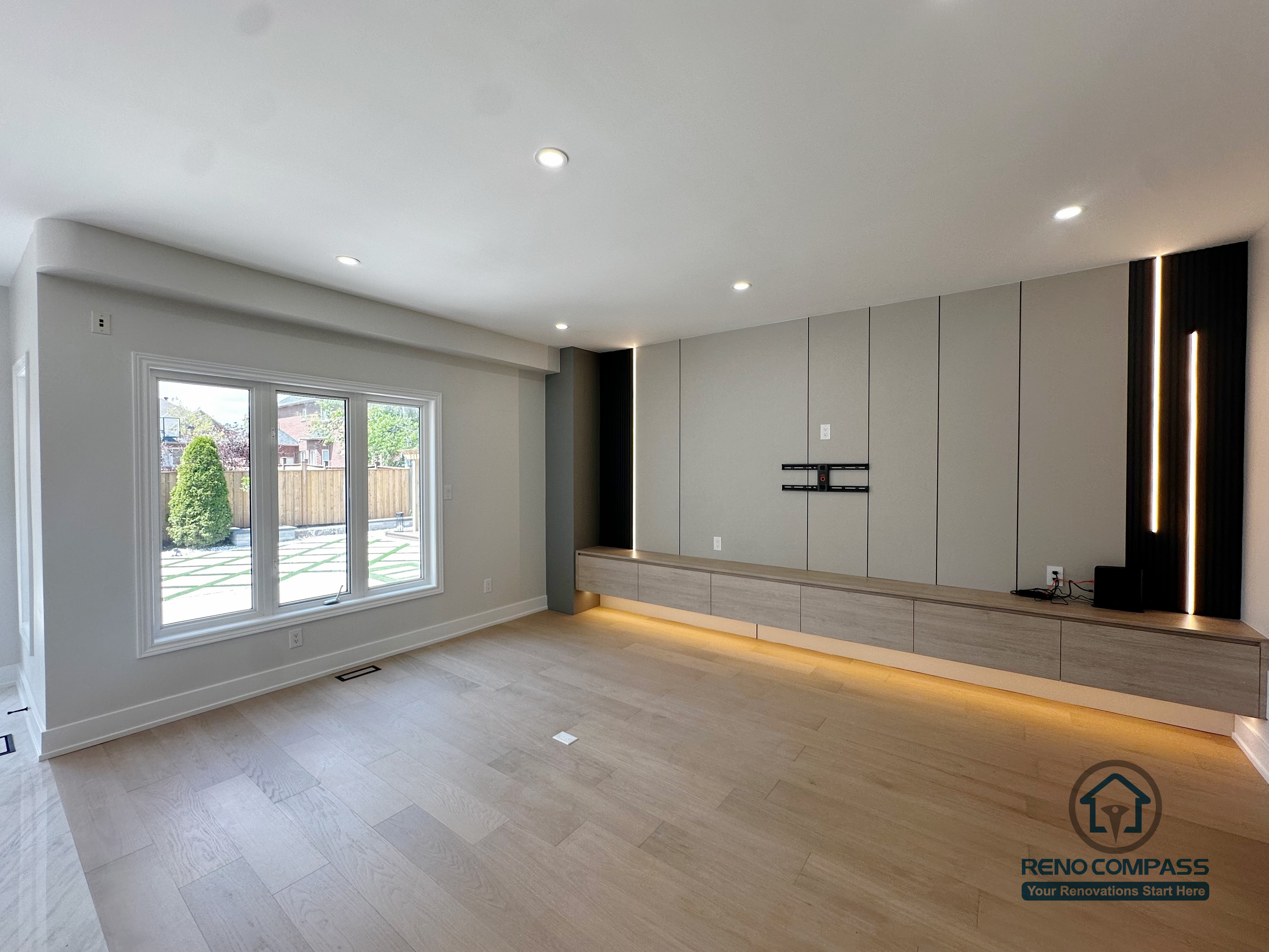 This image showcases a modern living room completed as part of a Markham interior renovation, featuring a custom-designed TV feature wall that balances clean lines with subtle lighting details. Full-height wall panels create a structured backdrop, while integrated vertical and base LED lighting adds warmth and depth without overwhelming the space.

A low-profile floating media cabinet runs along the wall, offering practical storage while keeping the floor visually open. The concealed wiring and wall-mounted TV setup ensure a clean, uncluttered appearance that supports both daily living and entertainment use.

Natural light from the large window enhances the neutral color palette and light wood flooring, reinforcing a calm and spacious atmosphere. This living room renovation reflects Reno Compass’s approach to interior design—where functionality, proportion, and lighting are carefully coordinated for modern Markham homes.