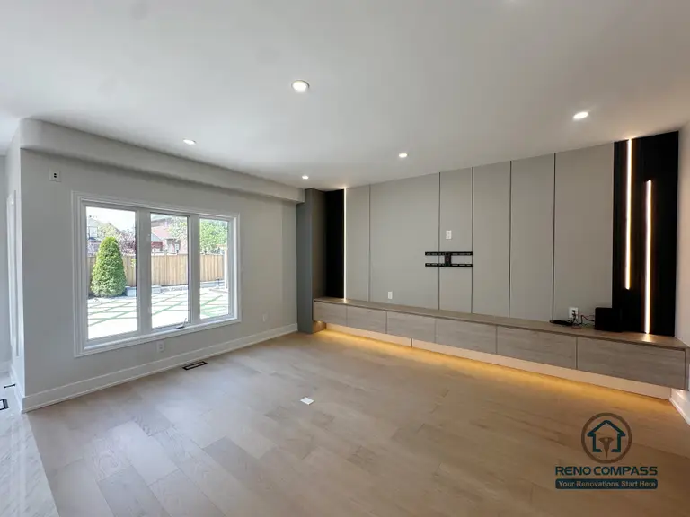 This image showcases a modern living room completed as part of a Markham interior renovation, featuring a custom-designed TV feature wall that balances clean lines with subtle lighting details. Full-height wall panels create a structured backdrop, while integrated vertical and base LED lighting adds warmth and depth without overwhelming the space.

A low-profile floating media cabinet runs along the wall, offering practical storage while keeping the floor visually open. The concealed wiring and wall-mounted TV setup ensure a clean, uncluttered appearance that supports both daily living and entertainment use.

Natural light from the large window enhances the neutral color palette and light wood flooring, reinforcing a calm and spacious atmosphere. This living room renovation reflects Reno Compass’s approach to interior design—where functionality, proportion, and lighting are carefully coordinated for modern Markham homes.