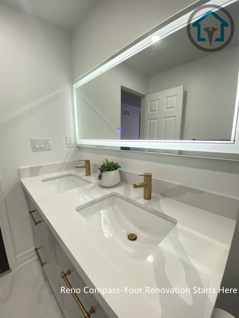 Close-up view of a modern double vanity installed as part of a Pickering bathroom renovation. The design features an integrated white quartz countertop with dual undermount sinks, brushed brass faucets, and a full-width LED backlit mirror. Clean lines, neutral tones, and balanced lighting create a functional yet refined vanity area suitable for daily family use.
