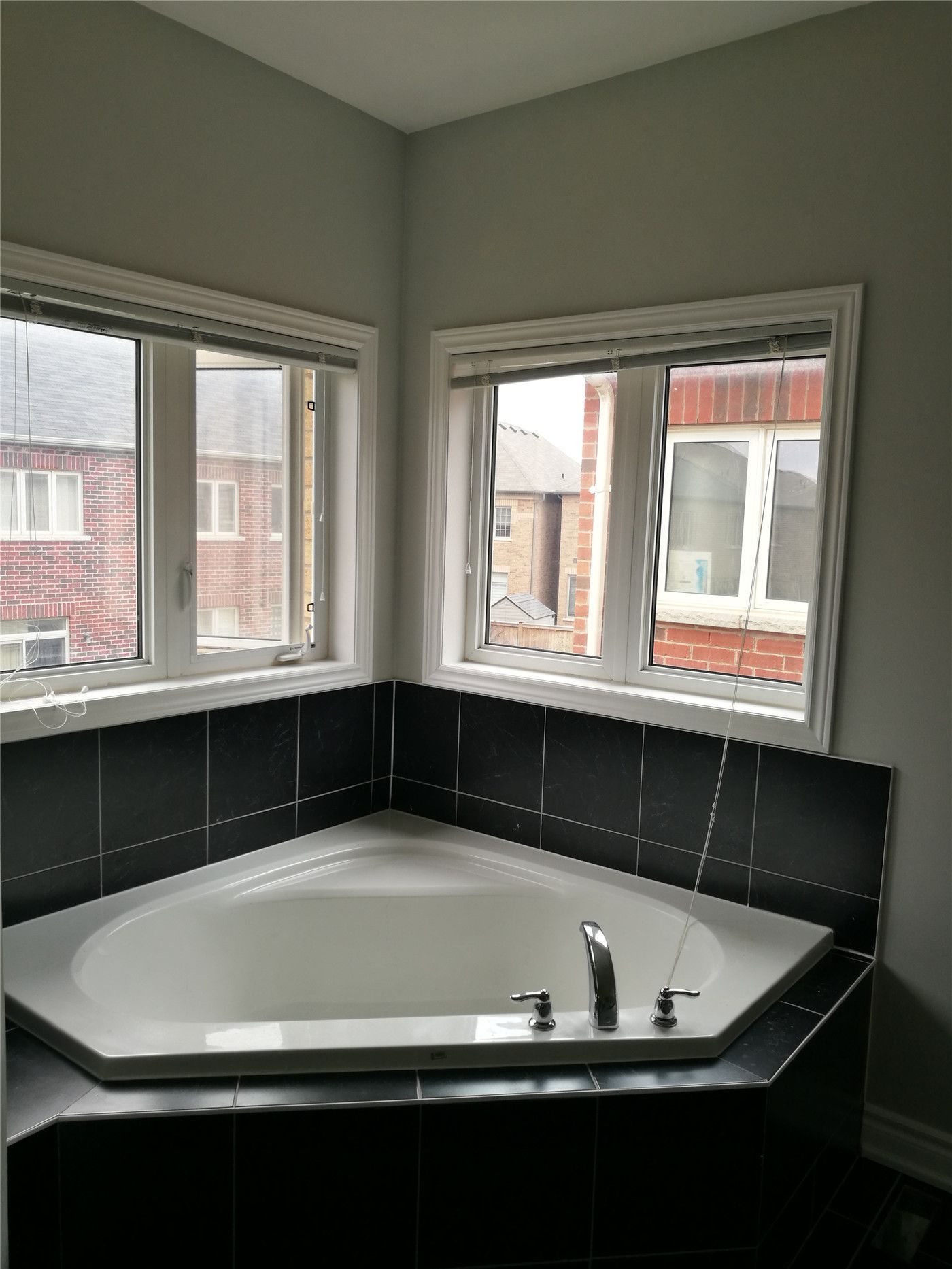 This image features a bathtub area after a Markham interior renovation, showcasing a corner soaking tub, tiled surround, upgraded fixtures, and dual windows that bring in natural light and improve the overall bathroom experience.