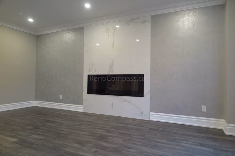 This Markham interior renovation highlights a modern living room upgrade featuring a sleek built-in linear fireplace and a marble-style feature wall. The space is finished with warm neutral paint tones, crown molding details, and newly installed dark hardwood flooring, creating a clean and elegant contemporary atmosphere. Completed by Reno Compass, a trusted renovation contractor providing professional interior renovation and custom finishing services in Markham and the Greater Toronto Area.