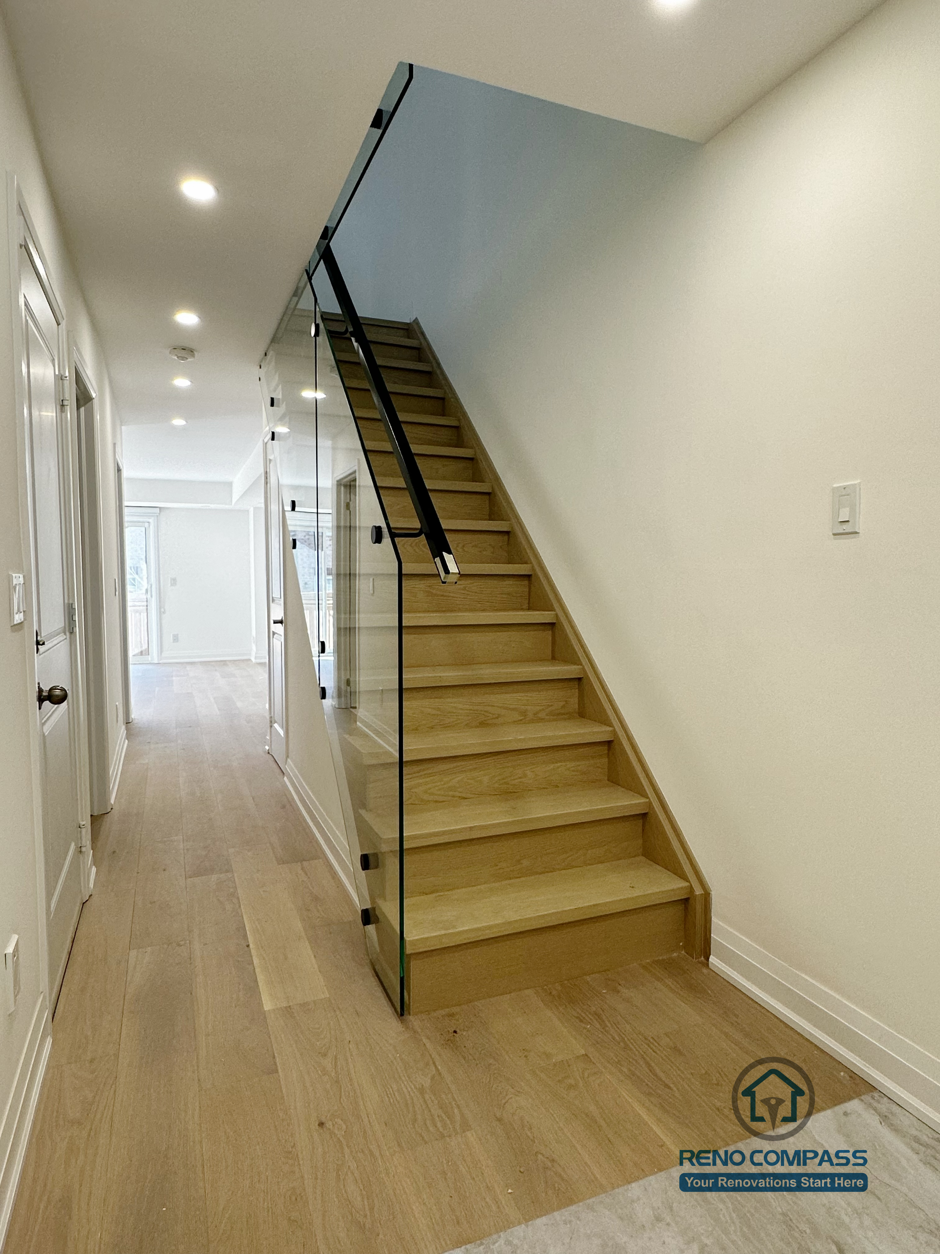 This Richmond Hill interior renovation features a modern staircase with natural oak treads and a full-height glass railing, creating a clean and open transition between floors. The minimalist design enhances light flow along the hallway, while the warm wood flooring and recessed lighting provide a cohesive, contemporary interior aesthetic.