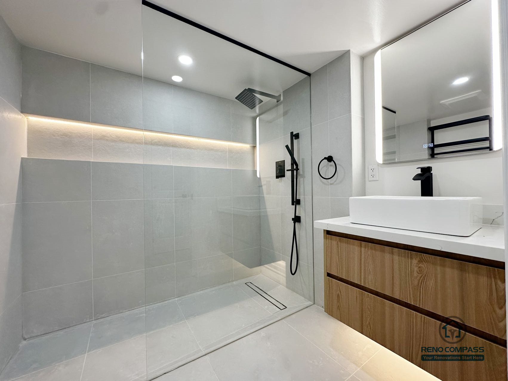 This image highlights a thoughtfully designed curbless walk-in shower with a linear floor drain and integrated niche lighting. The seamless floor transition, large-format wall tiles, and restrained color palette create a calm and balanced bathroom environment. Matte black fixtures contrast subtly with the light surfaces, while the floating vanity adds warmth and visual lightness. The layout prioritizes drainage efficiency, spatial clarity, and long-term daily comfort.