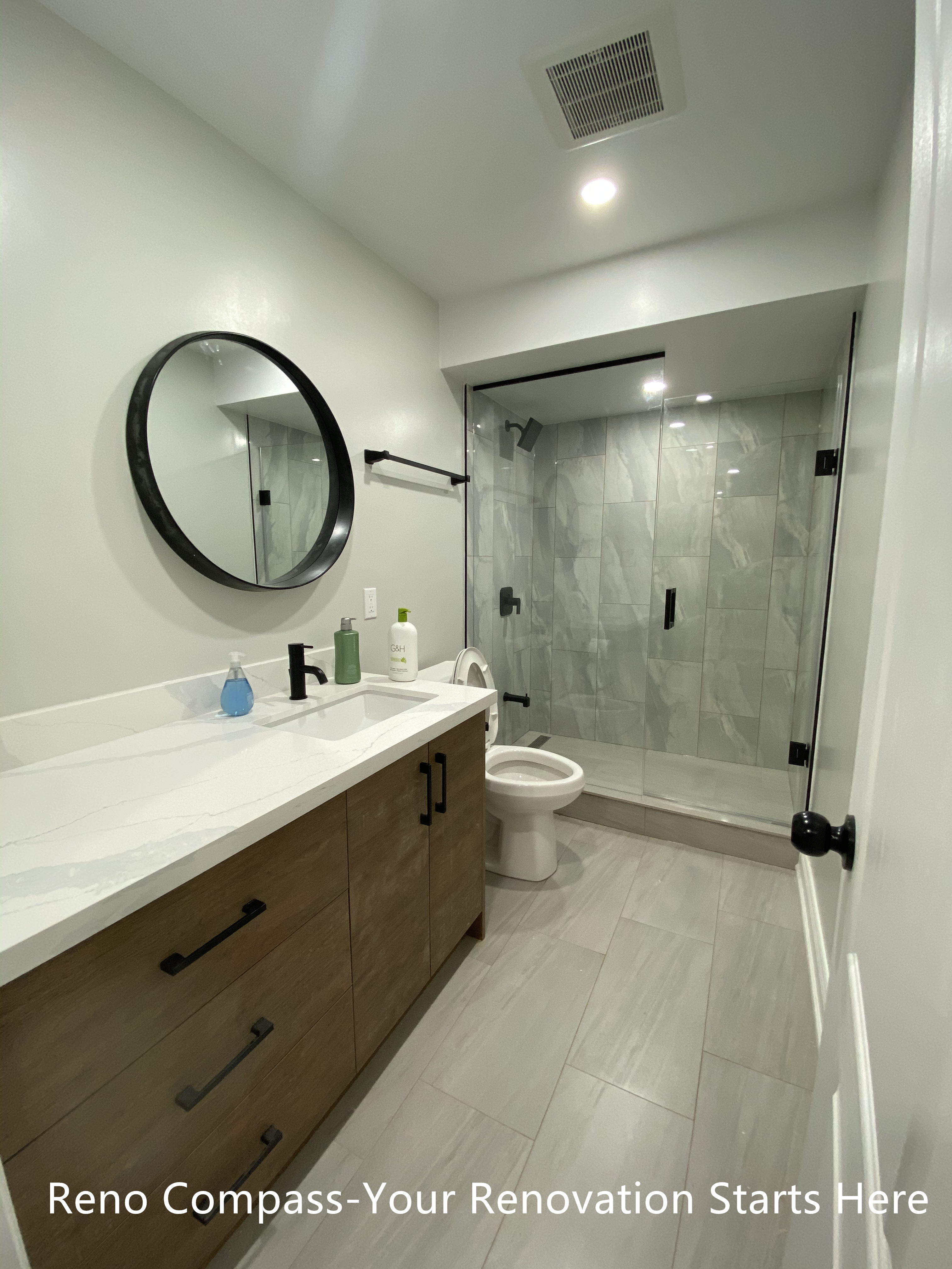 This Aurora basement bathroom renovation features a modern and space-efficient layout designed for daily use. A custom wood-tone vanity with integrated storage is paired with a quartz countertop and a round black-framed mirror, adding warmth and contrast to the neutral palette. The walk-in shower is finished with large-format porcelain tiles and a frameless glass enclosure, creating a clean and open visual effect while maximizing usable space. Thoughtful lighting and durable materials make this bathroom both practical and easy to maintain. Completed by Reno Compass, a Toronto renovation contractor specializing in basement renovations.