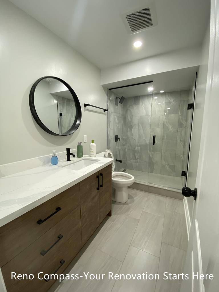 This Aurora basement bathroom renovation features a modern and space-efficient layout designed for daily use. A custom wood-tone vanity with integrated storage is paired with a quartz countertop and a round black-framed mirror, adding warmth and contrast to the neutral palette. The walk-in shower is finished with large-format porcelain tiles and a frameless glass enclosure, creating a clean and open visual effect while maximizing usable space. Thoughtful lighting and durable materials make this bathroom both practical and easy to maintain. Completed by Reno Compass, a Toronto renovation contractor specializing in basement renovations.
