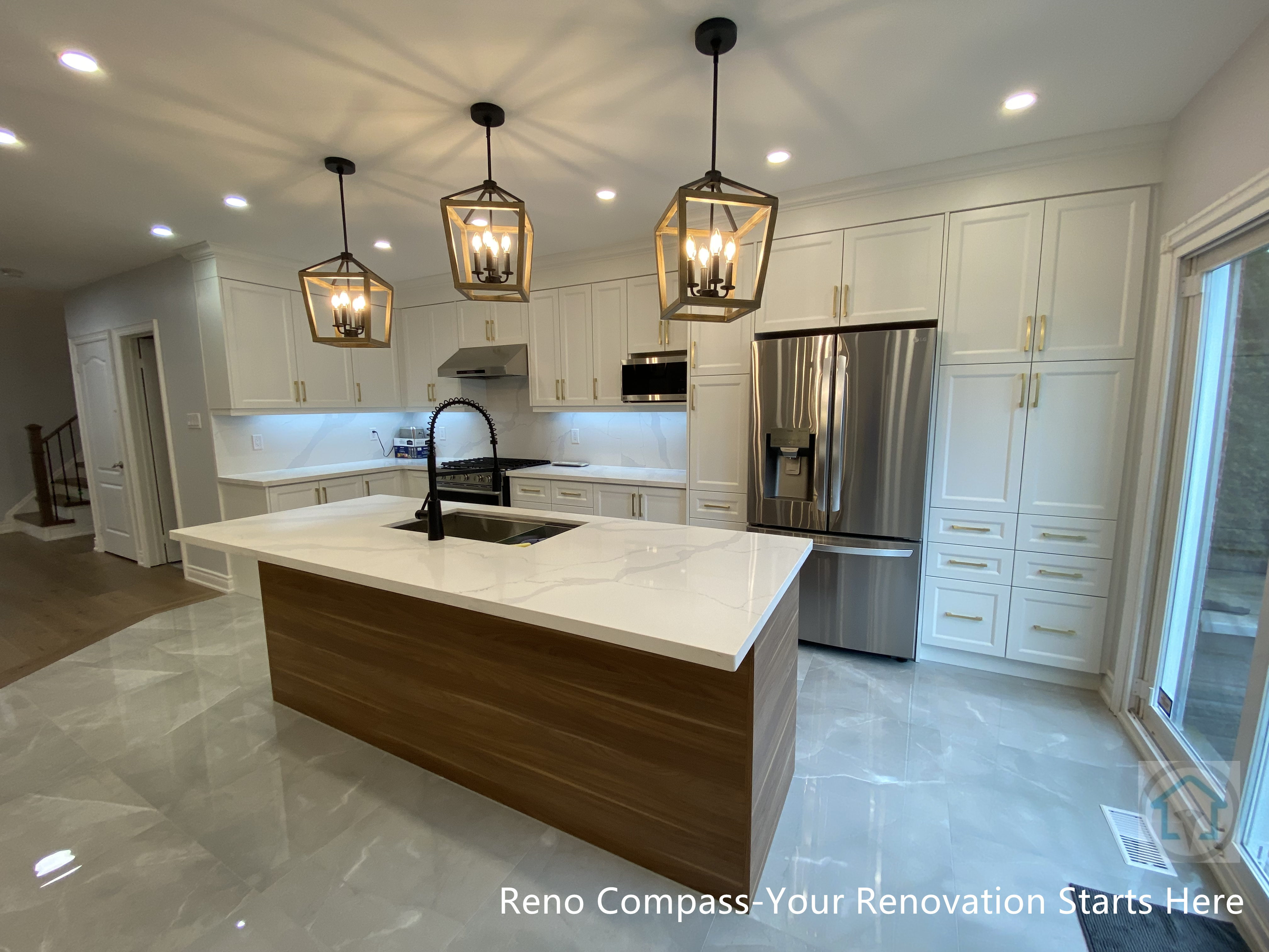 This image highlights a Newmarket interior renovation featuring a long quartz kitchen island with warm wood base cabinetry, designed to anchor the open-concept layout. The linear arrangement of lantern-style pendant lights creates strong visual rhythm and enhances the sense of depth within the space. White shaker cabinets, integrated stainless steel appliances, and glossy marble-look flooring work together to reflect light and elevate the overall brightness. A high-arc faucet and built-in sink add both functionality and modern appeal. Completed by a Toronto renovation company, this kitchen renovation emphasizes clean lines, balanced proportions, and seamless flow between cooking, dining, and outdoor access.