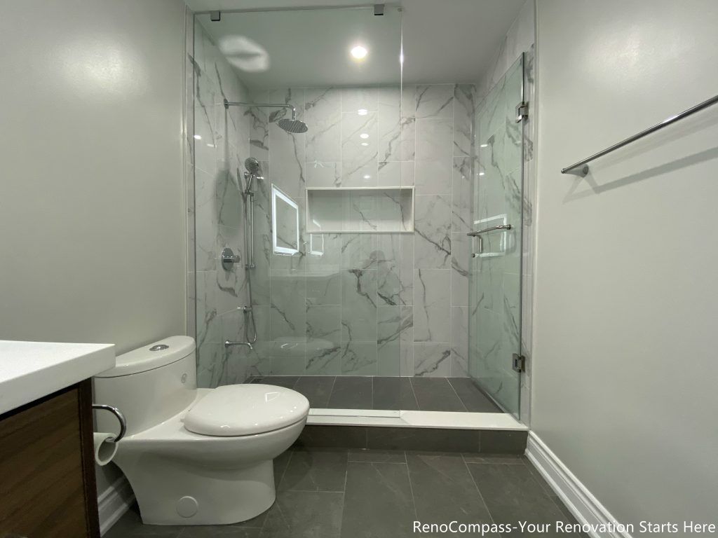 This Oakville bathroom renovation showcases a modern walk-in glass shower with marble-look wall tiles, built-in shower niche, and sleek frameless enclosure for a clean contemporary design. The space also features a custom wood vanity with quartz countertop, LED backlit mirror, and upgraded fixtures that enhance both functionality and visual appeal. Completed by a professional Toronto renovation company, this bathroom upgrade focuses on maximizing brightness, storage, and comfort while delivering a stylish and durable modern bathroom space.