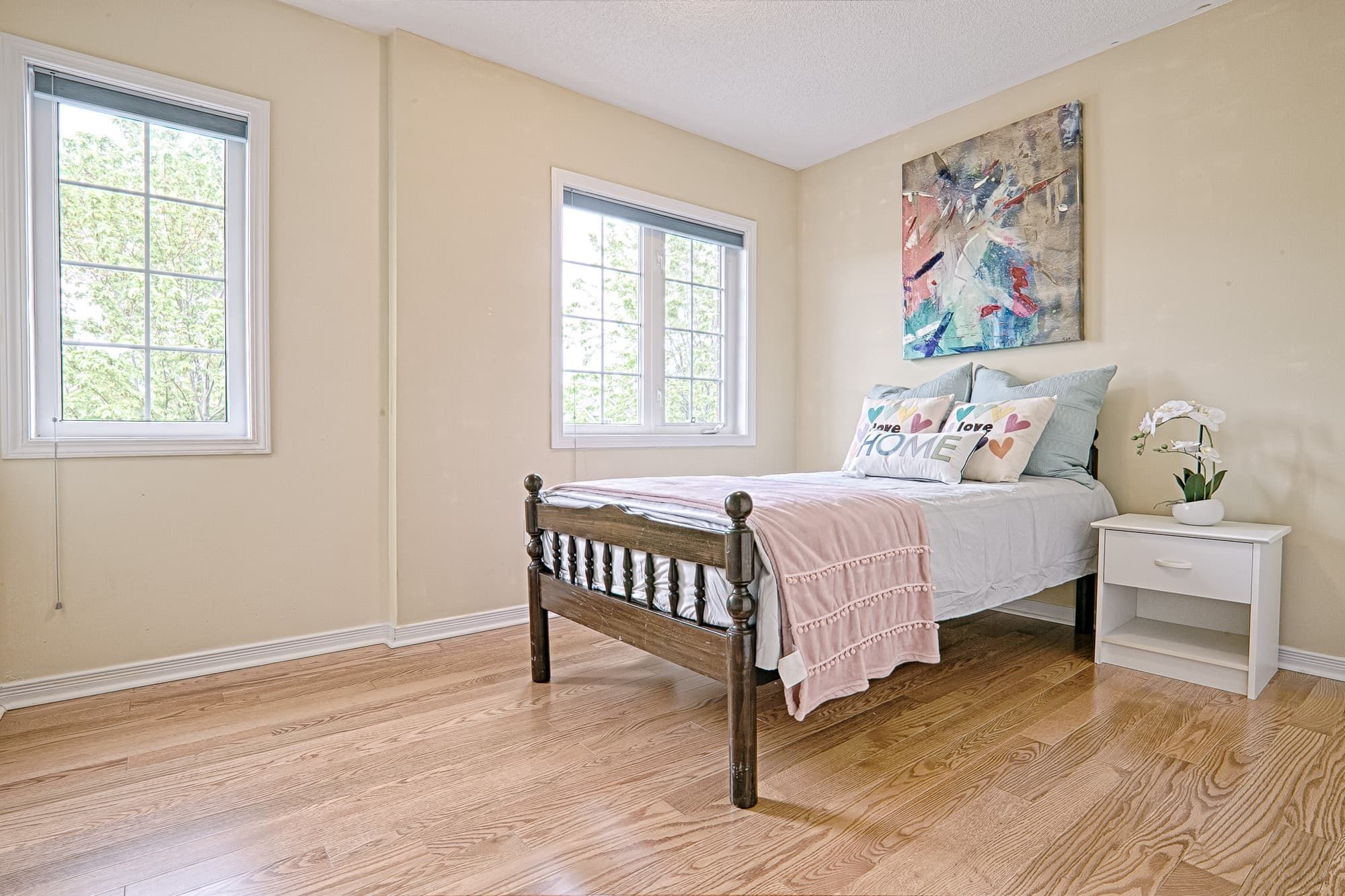 This cozy bedroom showcases a simple and lovely interior design with warm hardwood flooring, soft neutral wall colors, and abundant natural light from large windows. The wooden bed frame paired with light bedding creates a calm and comfortable atmosphere, while minimal decor enhances the sense of space. This design emphasizes practicality, brightness, and everyday comfort, making it ideal for modern family homes and rental properties.