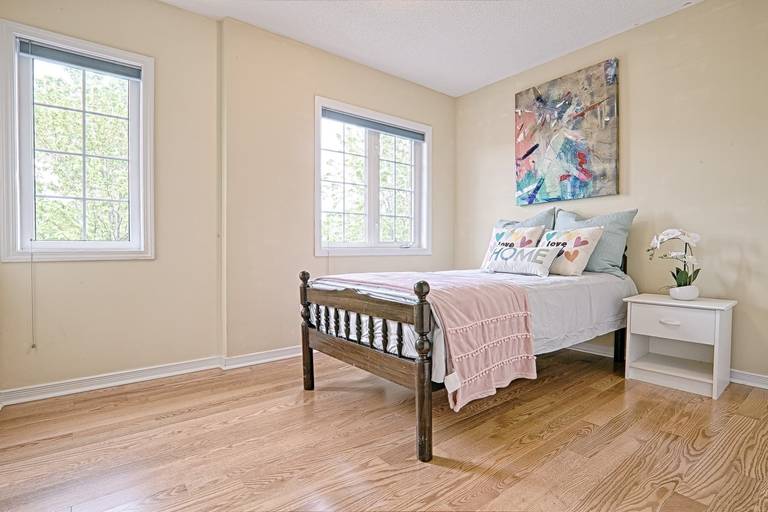 This cozy bedroom showcases a simple and lovely interior design with warm hardwood flooring, soft neutral wall colors, and abundant natural light from large windows. The wooden bed frame paired with light bedding creates a calm and comfortable atmosphere, while minimal decor enhances the sense of space. This design emphasizes practicality, brightness, and everyday comfort, making it ideal for modern family homes and rental properties.