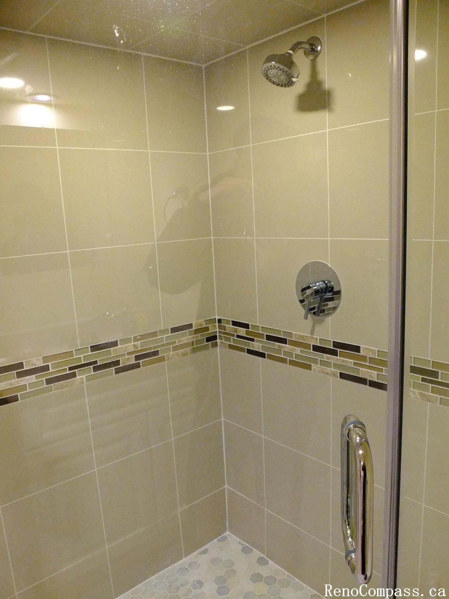 This Mississauga bathroom renovation showcases a modern walk-in shower featuring clean beige wall tiles and a decorative mosaic accent band that adds visual depth to the space. The shower area is equipped with a chrome shower fixture, frameless glass enclosure, and a pebble-style shower floor designed for both comfort and slip resistance. The neutral color palette creates a warm and inviting atmosphere while maintaining a timeless look. This renovation highlights quality tile installation and thoughtful design that improves both functionality and aesthetics in a modern bathroom space.