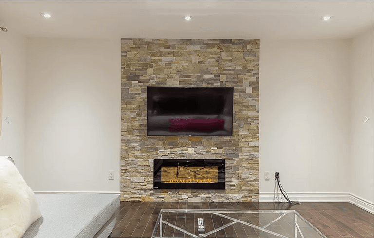 A beautifully renovated living room from our Vaughan full house interior project featuring a natural stone feature wall with an electric fireplace and wall-mounted TV. The warm tones, recessed lighting, and minimalist design create a cozy yet modern atmosphere perfect for family living.
