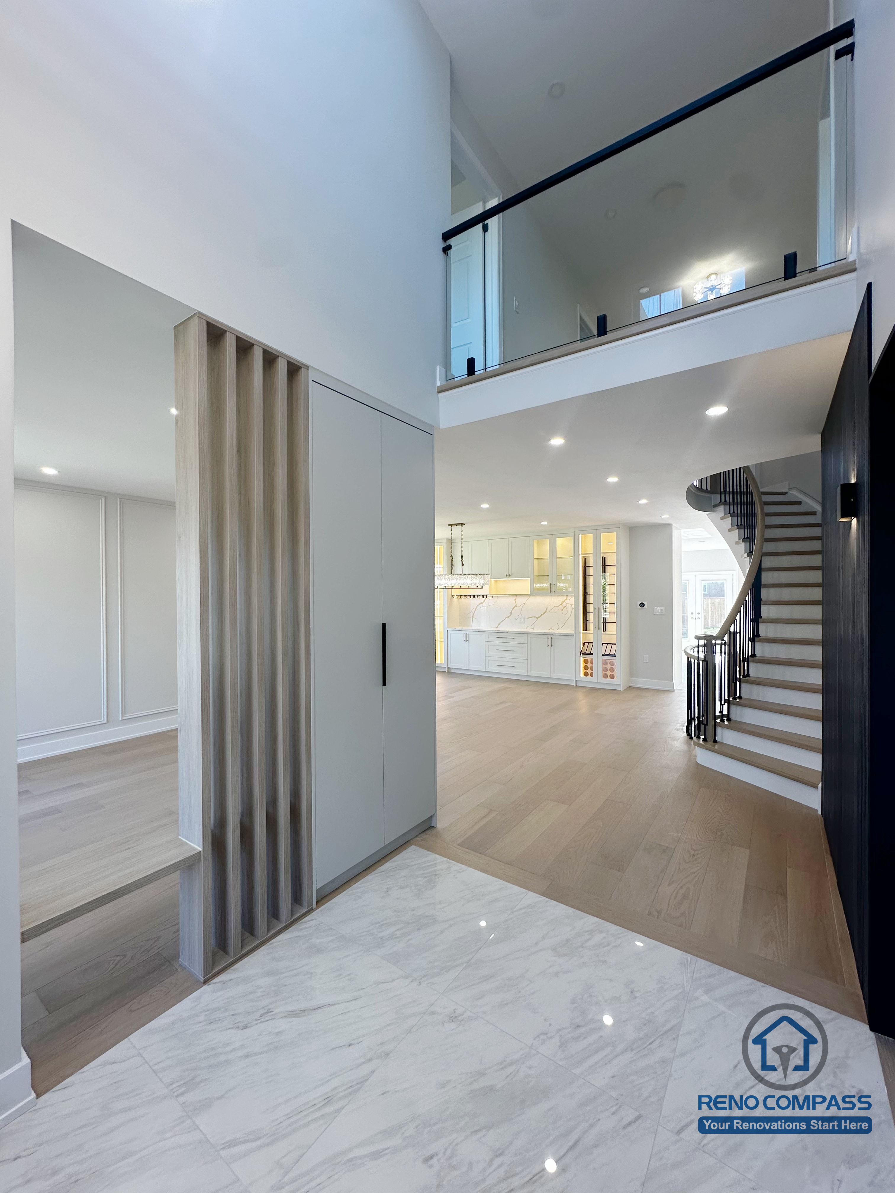This Markham interior renovation showcases a double-height foyer designed to enhance openness, natural light, and visual flow throughout the home. A custom vertical slat feature subtly defines adjacent spaces without blocking sightlines, while the curved staircase and glass railing add architectural movement and elegance. The combination of marble-look flooring, warm wood finishes, and recessed lighting creates a balanced transition between entry, living areas, and the kitchen beyond. The result is a refined interior layout that feels open, structured, and thoughtfully connected.