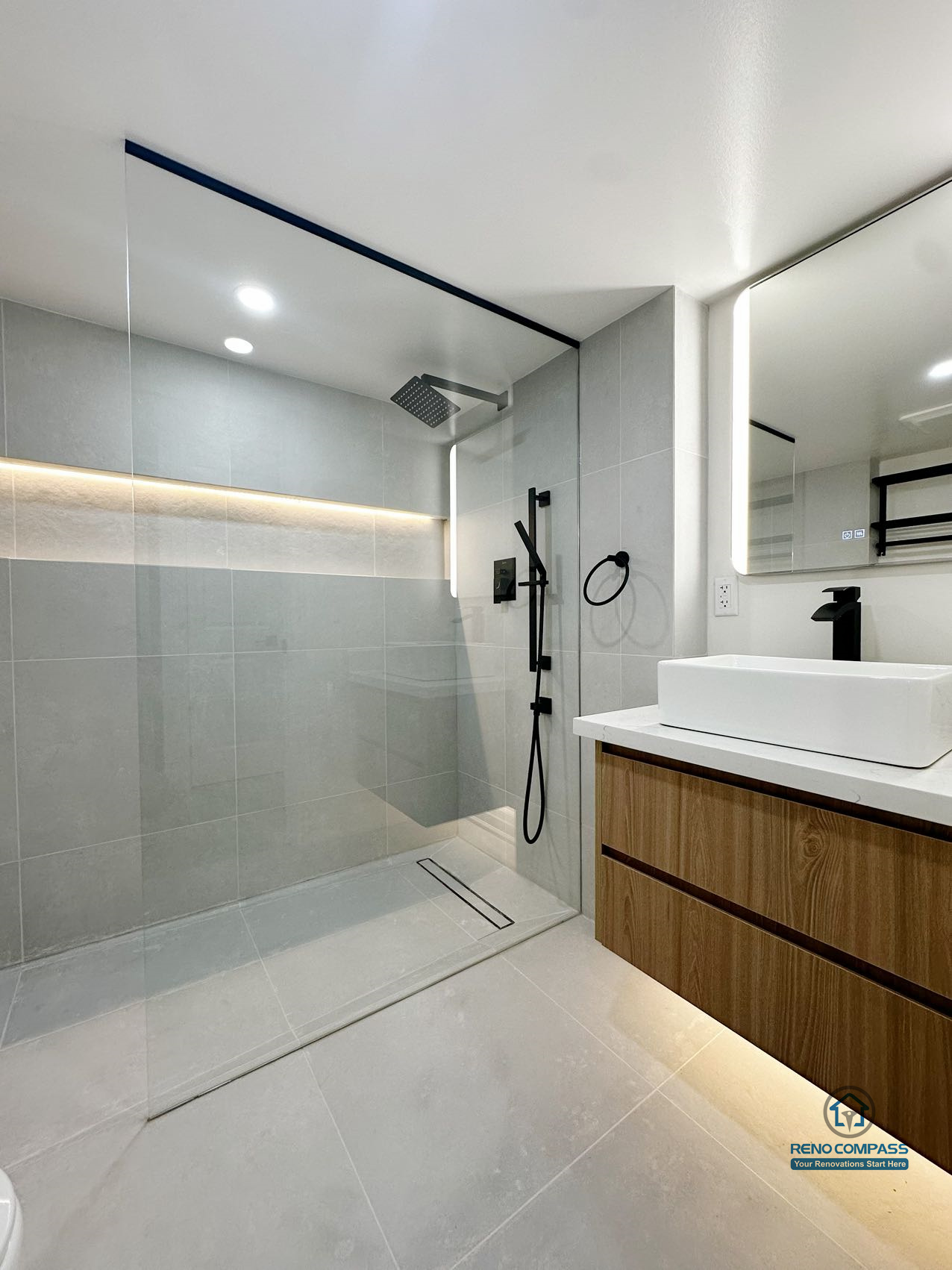 This image highlights a thoughtfully designed curbless walk-in shower with a linear floor drain and integrated niche lighting. The seamless floor transition, large-format wall tiles, and restrained color palette create a calm and balanced bathroom environment. Matte black fixtures contrast subtly with the light surfaces, while the floating vanity adds warmth and visual lightness. The layout prioritizes drainage efficiency, spatial clarity, and long-term daily comfort.