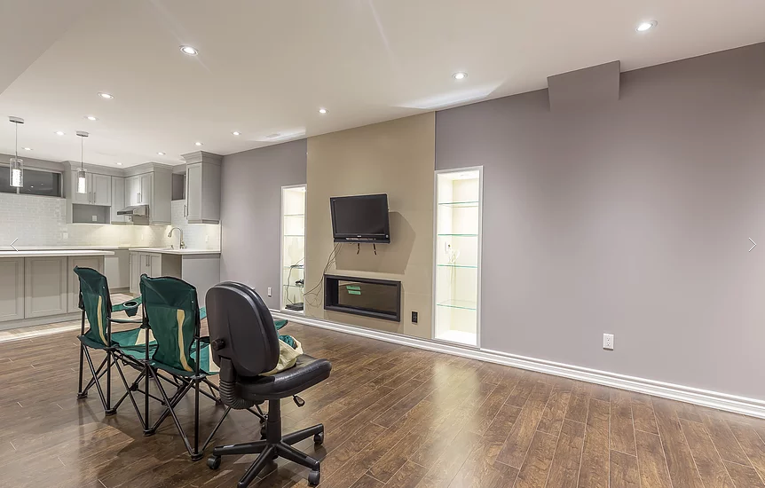 This Toronto basement renovation showcases a modern open-concept entertainment space featuring a built-in electric fireplace, TV feature wall, custom glass display niches, and upgraded hardwood flooring. The basement also includes a fully finished kitchen with quartz countertops, grey cabinetry, and recessed pot lighting. Designed for comfortable family living and entertaining, this project highlights the quality craftsmanship of a professional Toronto renovation company specializing in basement finishing.