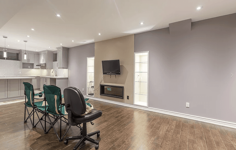 This Toronto basement renovation showcases a modern open-concept entertainment space featuring a built-in electric fireplace, TV feature wall, custom glass display niches, and upgraded hardwood flooring. The basement also includes a fully finished kitchen with quartz countertops, grey cabinetry, and recessed pot lighting. Designed for comfortable family living and entertaining, this project highlights the quality craftsmanship of a professional Toronto renovation company specializing in basement finishing.