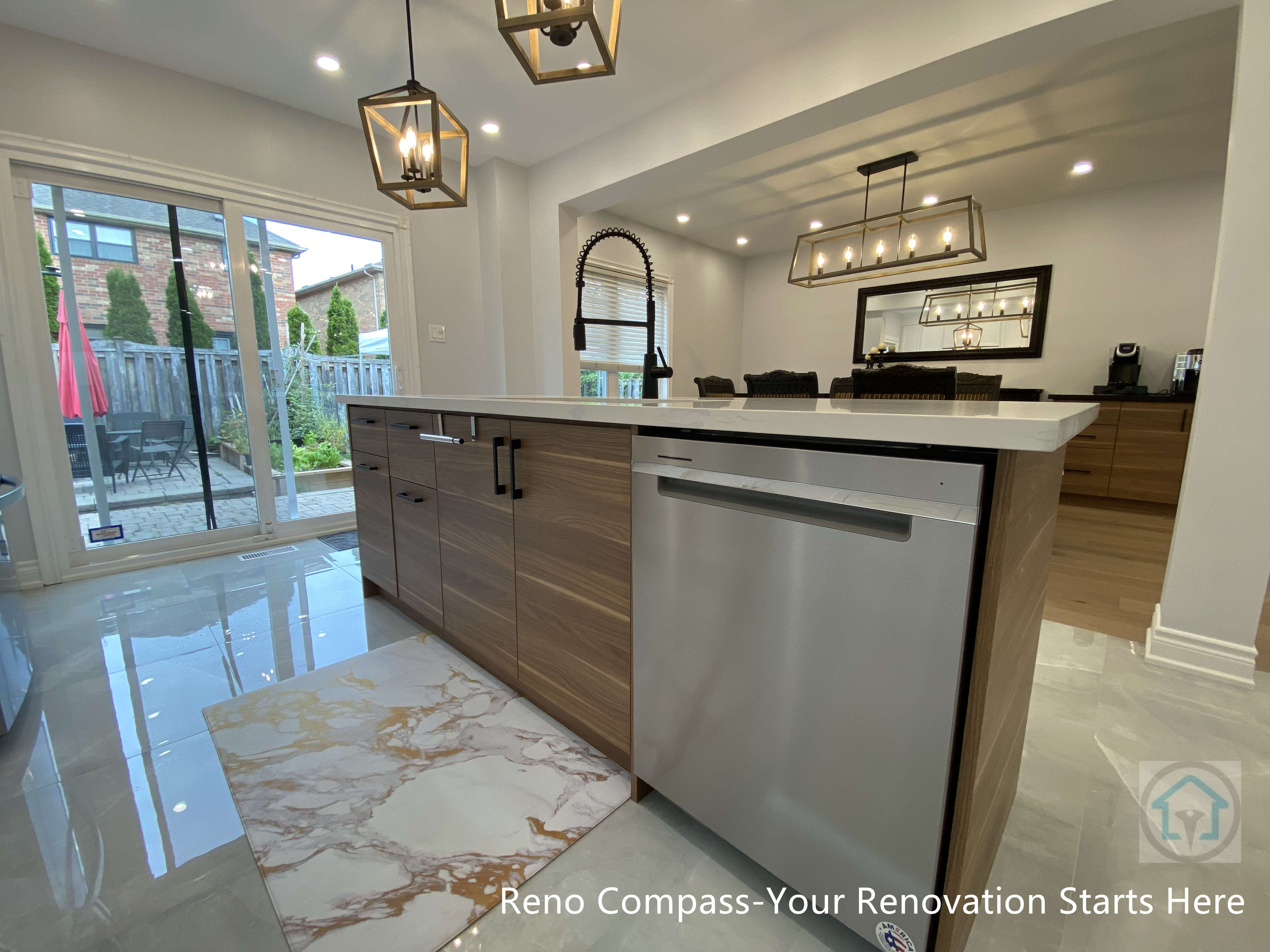 Modern interior renovation in Newmarket featuring a custom-built kitchen island with integrated dishwasher and warm wood cabinetry. The polished tile flooring reflects natural light from the sliding patio doors, enhancing brightness and spatial depth. Black hardware, statement pendant lighting, and a streamlined faucet add contrast and visual focus. This open-concept kitchen seamlessly connects to the dining area, demonstrating thoughtful layout planning and craftsmanship by a Toronto renovation company specializing in full home interior renovations.