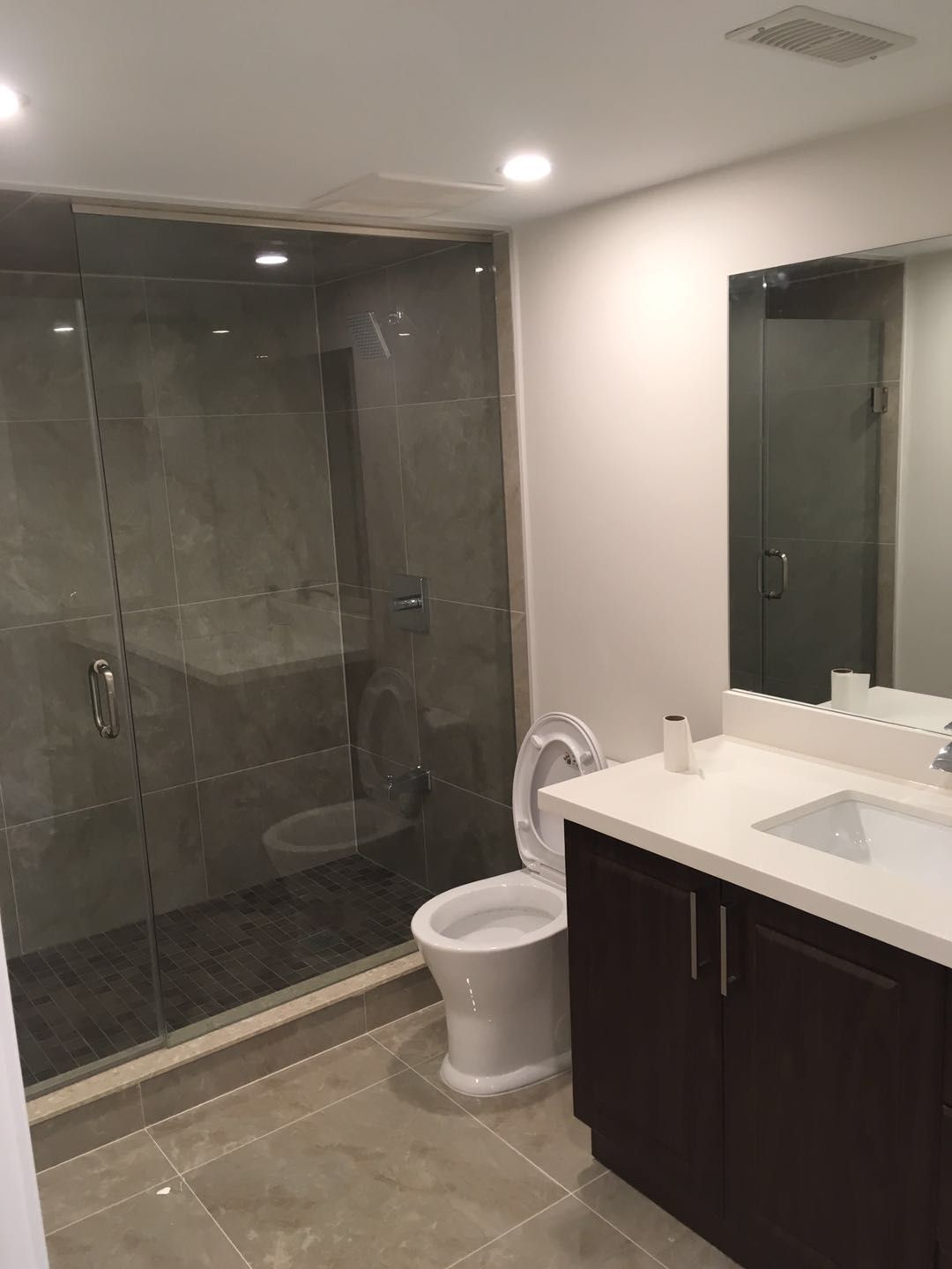 This Toronto bathroom renovation features a spacious glass-enclosed shower with large-format wall tiles and a built-in bench for added comfort. The design includes a modern vanity with quartz countertop, a clean white toilet, and neutral tile flooring that enhances durability and ease of maintenance. Recessed lighting and a frameless mirror create a bright, functional, and contemporary bathroom space.