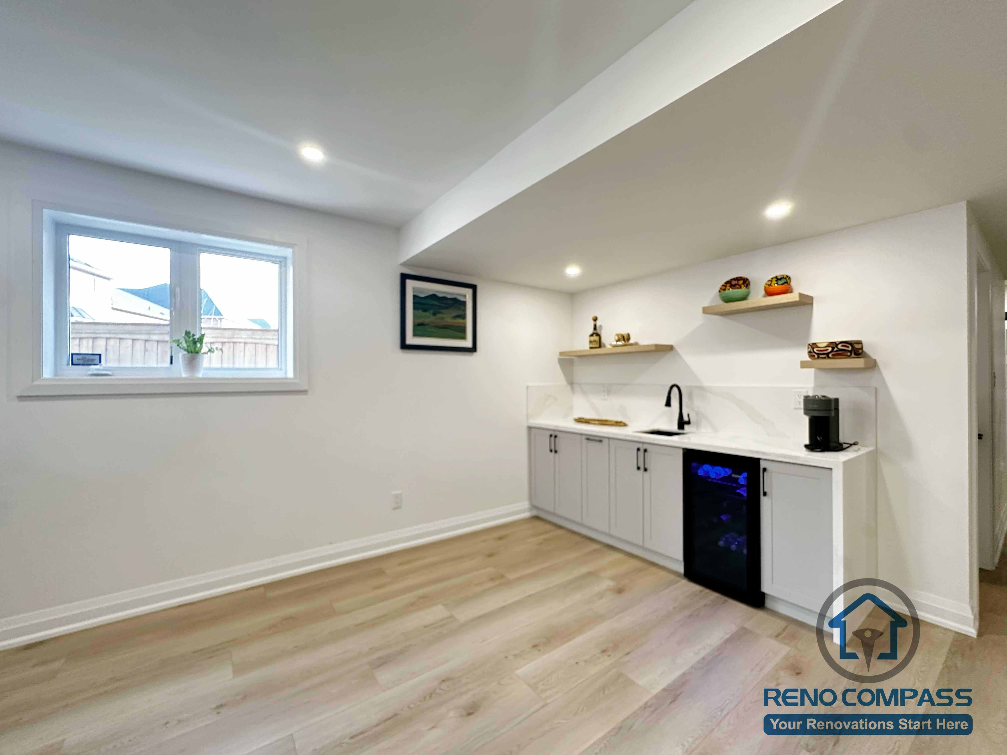 This image highlights a custom basement wet bar paired with an open multi-purpose area in a Richmond Hill basement renovation. The built-in cabinetry, floating shelves, sink, and integrated beverage fridge create a clean and functional bar setup, while the open floor space allows flexibility for a dining nook, lounge seating, or fitness area. Recessed lighting, wide-plank flooring, and natural light from the window help keep the basement bright and adaptable for everyday living or entertaining.