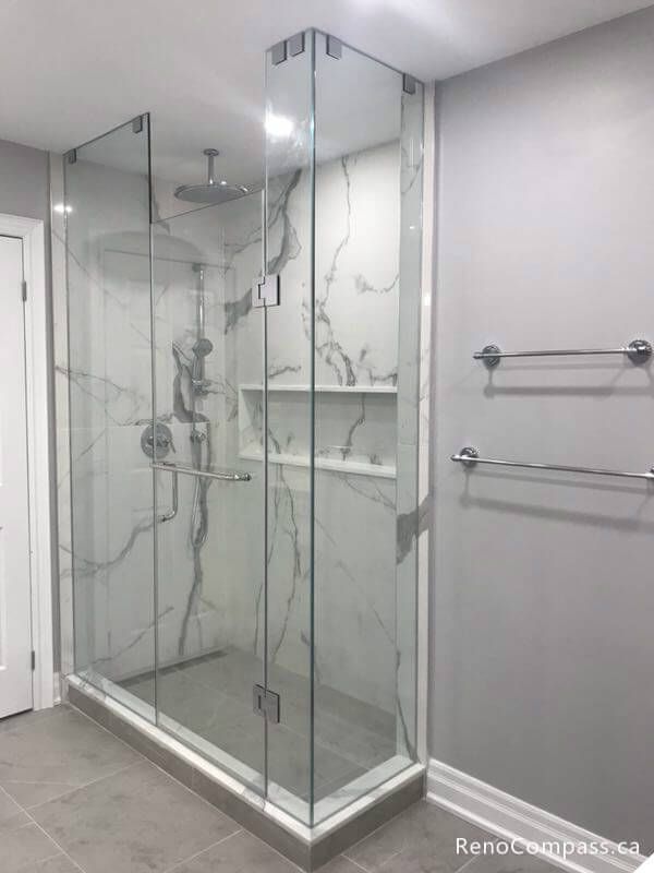 A luxury walk-in shower renovation in Markham featuring frameless glass doors, large-format marble-patterned wall tiles, a built-in shampoo niche, and a sleek rainfall showerhead.