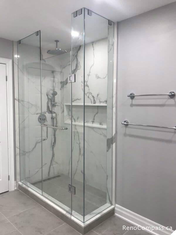 A luxury walk-in shower renovation in Markham featuring frameless glass doors, large-format marble-patterned wall tiles, a built-in shampoo niche, and a sleek rainfall showerhead.