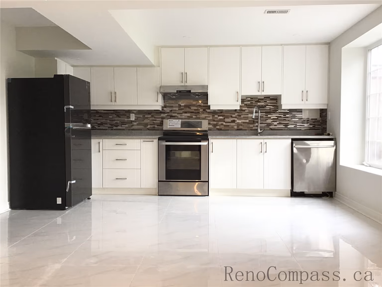 This Whitby basement renovation features a fully finished modern kitchen with sleek white cabinetry, stainless steel appliances, and a contemporary mosaic tile backsplash. The open layout, large window for natural light, and glossy porcelain flooring create a bright and functional secondary kitchen space — perfect for an in-law suite or basement apartment setup.