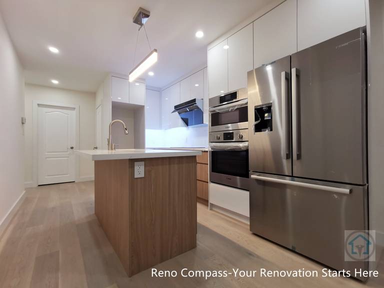 This image showcases a completed modern kitchen renovation in the Toronto area, featuring a clean island-centered layout with built-in stainless steel appliances. The design includes full-height white cabinetry, integrated oven and refrigerator units, wood-accent island panels, and recessed ceiling lighting. The open-concept layout improves circulation and functionality while maintaining a bright, minimal aesthetic. Completed by a professional Toronto renovation company, this kitchen renovation balances storage efficiency, modern finishes, and everyday usability for contemporary living.