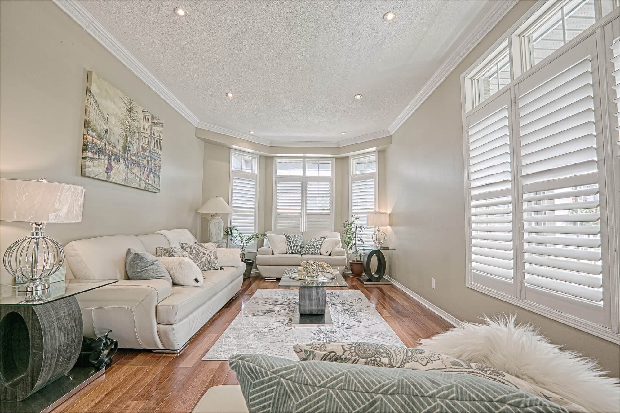 This elegant living room interior showcases warm hardwood flooring, soft neutral wall tones, and coordinated modern furniture that create a bright and sophisticated atmosphere. The large bay windows with plantation shutters allow abundant natural light, while the contemporary coffee table, textured area rug, and layered lighting enhance comfort and style. A refined example of warm minimalist living room renovation and modern home interior design inspiration for homeowners in Toronto and the GTA.
