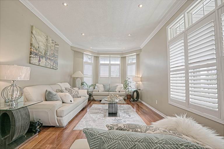This elegant living room interior showcases warm hardwood flooring, soft neutral wall tones, and coordinated modern furniture that create a bright and sophisticated atmosphere. The large bay windows with plantation shutters allow abundant natural light, while the contemporary coffee table, textured area rug, and layered lighting enhance comfort and style. A refined example of warm minimalist living room renovation and modern home interior design inspiration for homeowners in Toronto and the GTA.
