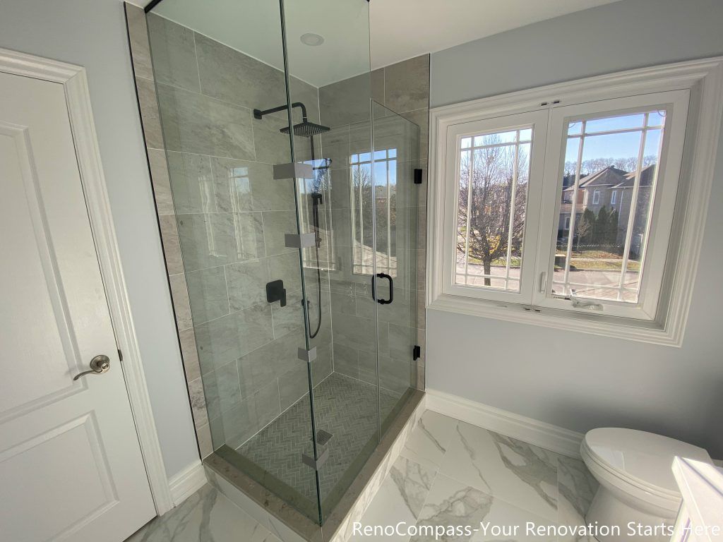 This Thornhill bathroom renovation features a frameless glass walk-in shower positioned beside a window, allowing natural light to enhance brightness and visual openness. Large-format gray wall tiles and a herringbone-patterned shower base create a clean, balanced look, while matte black fixtures add modern contrast. The transparent enclosure improves spatial flow and functionality, delivering a practical and well-lit bathroom designed for long-term everyday use.