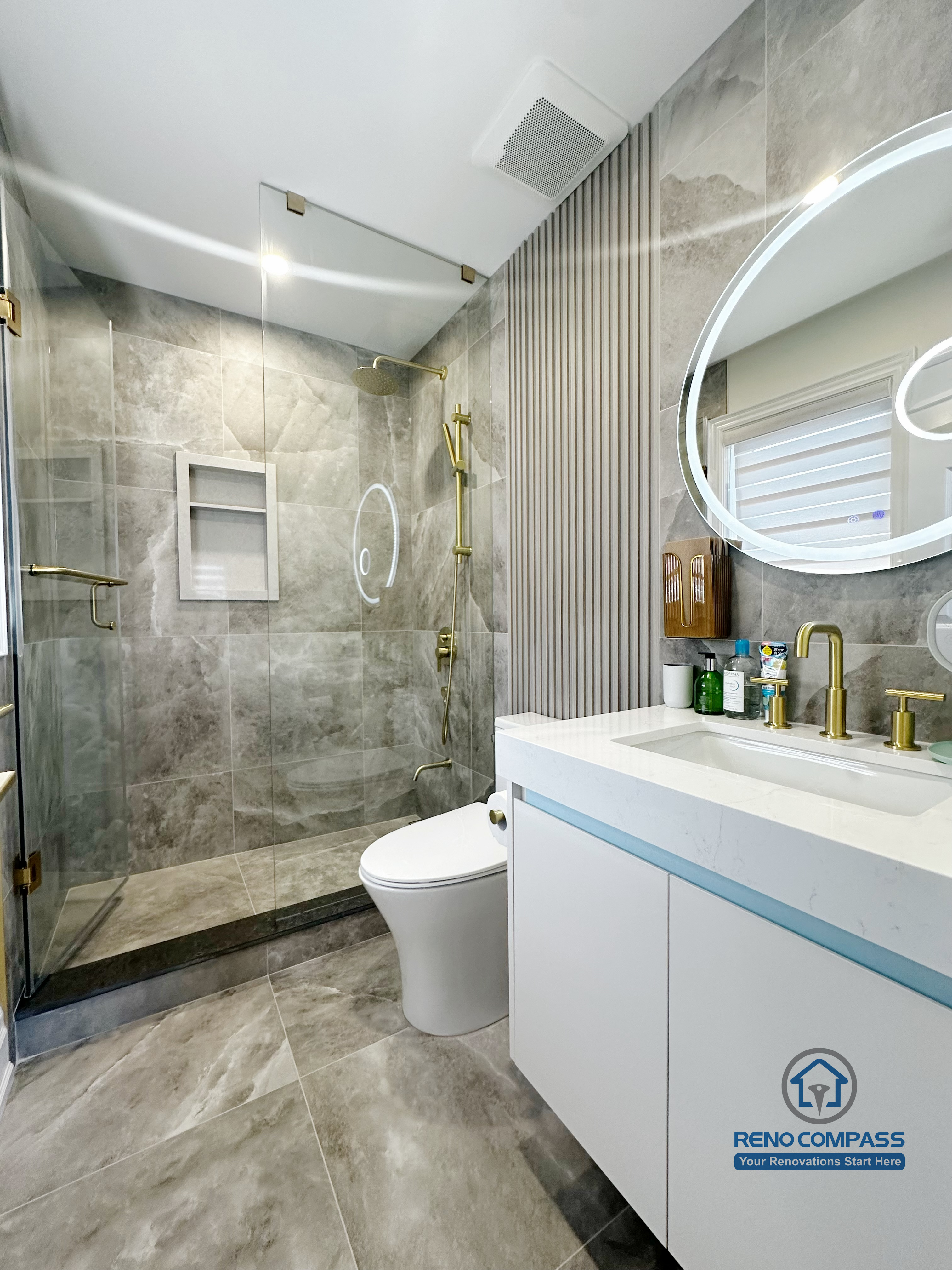This image showcases a modern bathroom renovation in North York, featuring a full-height glass shower enclosure with large-format grey stone tiles. Brushed gold shower fixtures and built-in wall niches add both function and visual contrast, while the floating vanity with integrated sink enhances the clean, contemporary look. The combination of neutral stone surfaces, reflective glass, and warm metal accents creates a refined and cohesive bathroom design focused on durability and everyday comfort.