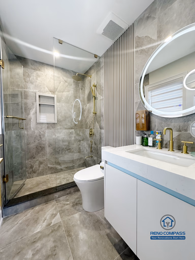 This image showcases a modern bathroom renovation in North York, featuring a full-height glass shower enclosure with large-format grey stone tiles. Brushed gold shower fixtures and built-in wall niches add both function and visual contrast, while the floating vanity with integrated sink enhances the clean, contemporary look. The combination of neutral stone surfaces, reflective glass, and warm metal accents creates a refined and cohesive bathroom design focused on durability and everyday comfort.