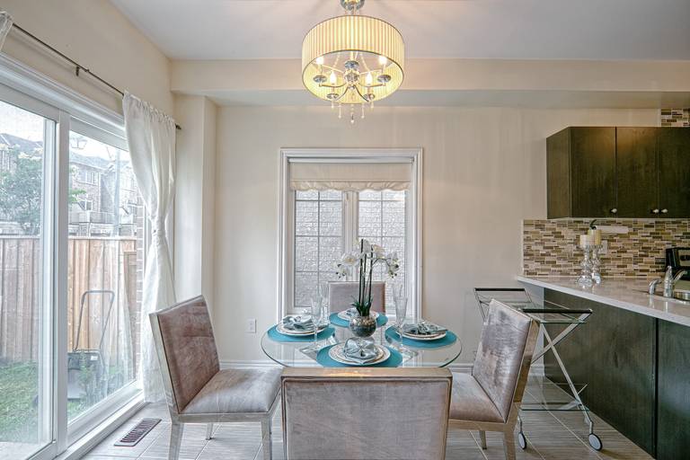 This elegant dining space features a modern glass-top dining table, upholstered chairs, and a stylish chandelier that enhances the warm and inviting atmosphere. Large sliding doors allow abundant natural light to fill the space, while the open connection to the contemporary kitchen with mosaic backsplash and dark cabinetry creates a cohesive and functional layout. Designed with both comfort and visual balance in mind, this interior reflects the quality craftsmanship of a professional Toronto renovation company specializing in modern residential upgrades and open-concept living design.