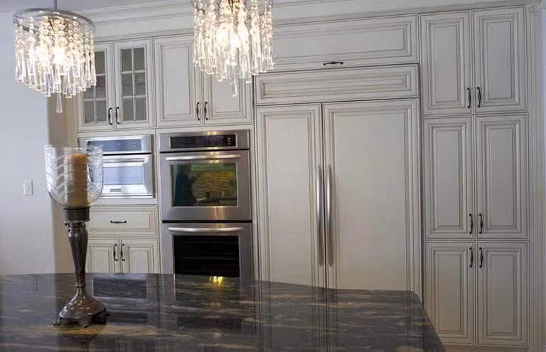 This European-style kitchen renovation showcases elegant custom white cabinetry with detailed panel molding and classic hardware, creating a refined and timeless aesthetic. The kitchen features a fully integrated built-in refrigerator and double wall ovens, maximizing functionality while maintaining a seamless design.

The large central island is finished with a dark natural stone countertop, adding contrast and sophistication to the space. Crystal chandeliers enhance the luxurious European atmosphere, combining traditional charm with modern convenience.

This kitchen design is ideal for homeowners seeking a high-end, classic European kitchen upgrade with enhanced storage, premium finishes, and a cohesive layout.