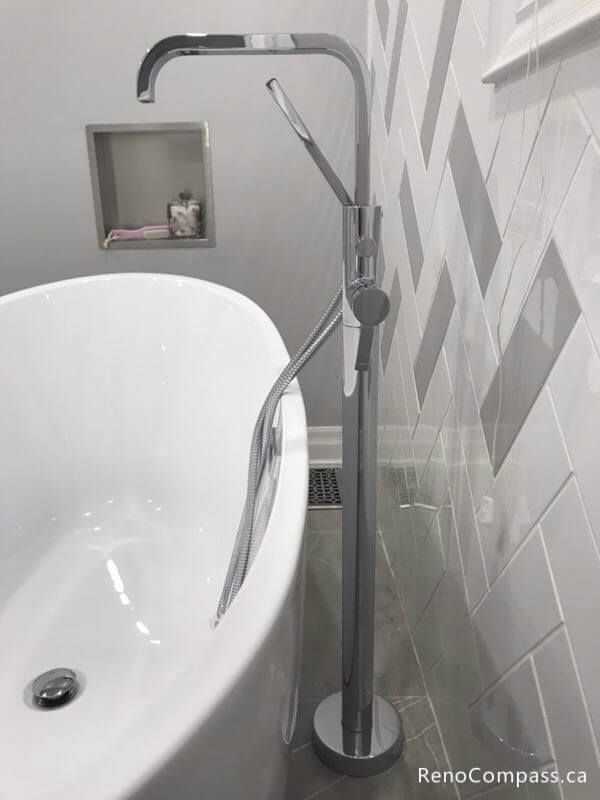 Close-up of a modern chrome floor-mount faucet paired with a sleek white freestanding bathtub. This Markham renovation features custom grey herringbone wall tiles and a built-in storage niche.