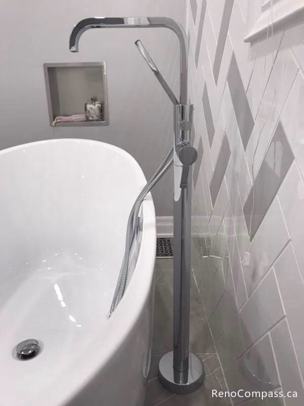 Close-up of a modern chrome floor-mount faucet paired with a sleek white freestanding bathtub. This Markham renovation features custom grey herringbone wall tiles and a built-in storage niche.