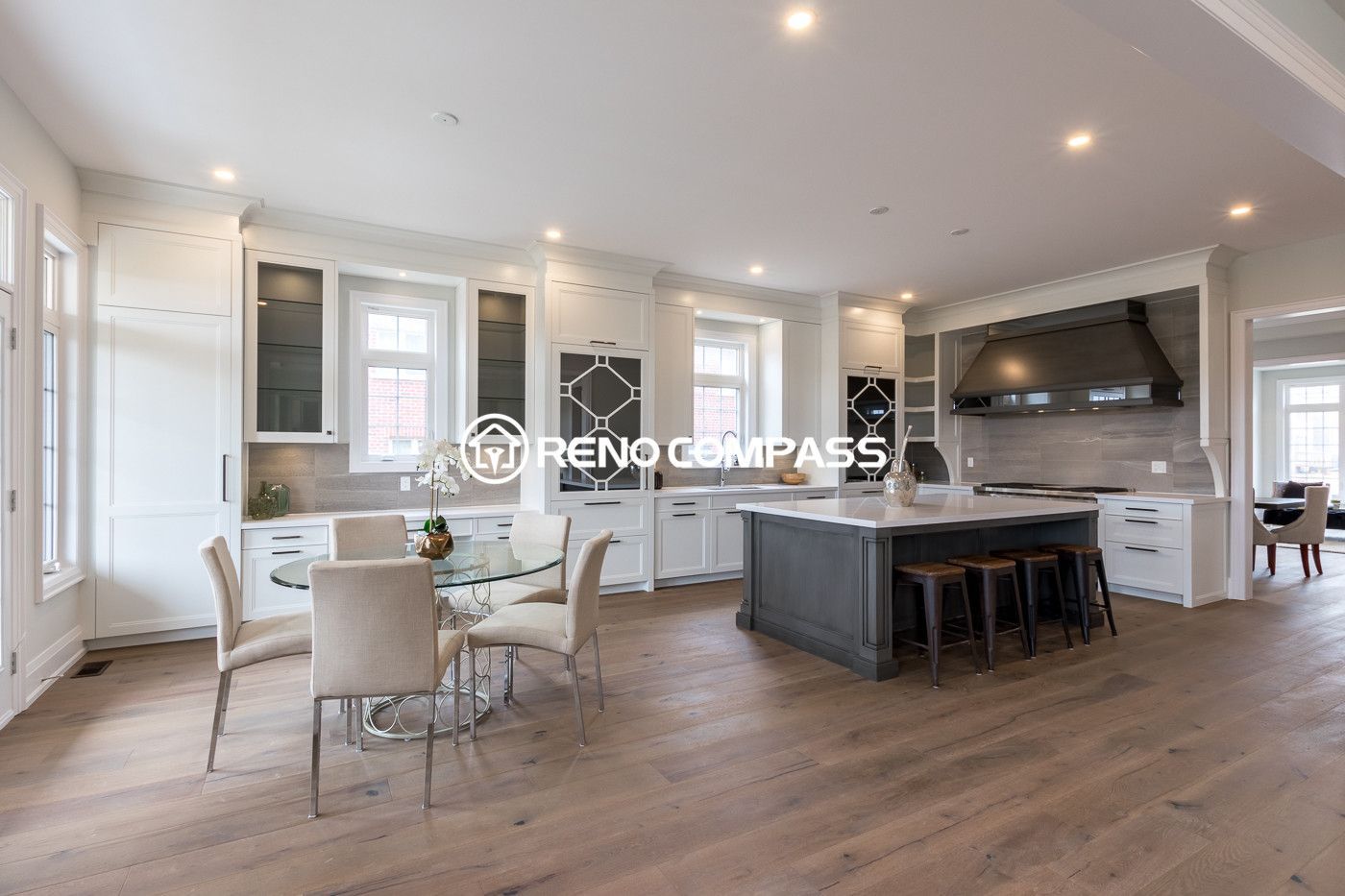 Bright open-concept kitchen and dining area renovation in Newmarket featuring custom white cabinetry, large windows, modern glass dining table, and engineered hardwood flooring. The design creates a spacious and elegant living environment that seamlessly connects the kitchen, dining, and outdoor access areas.