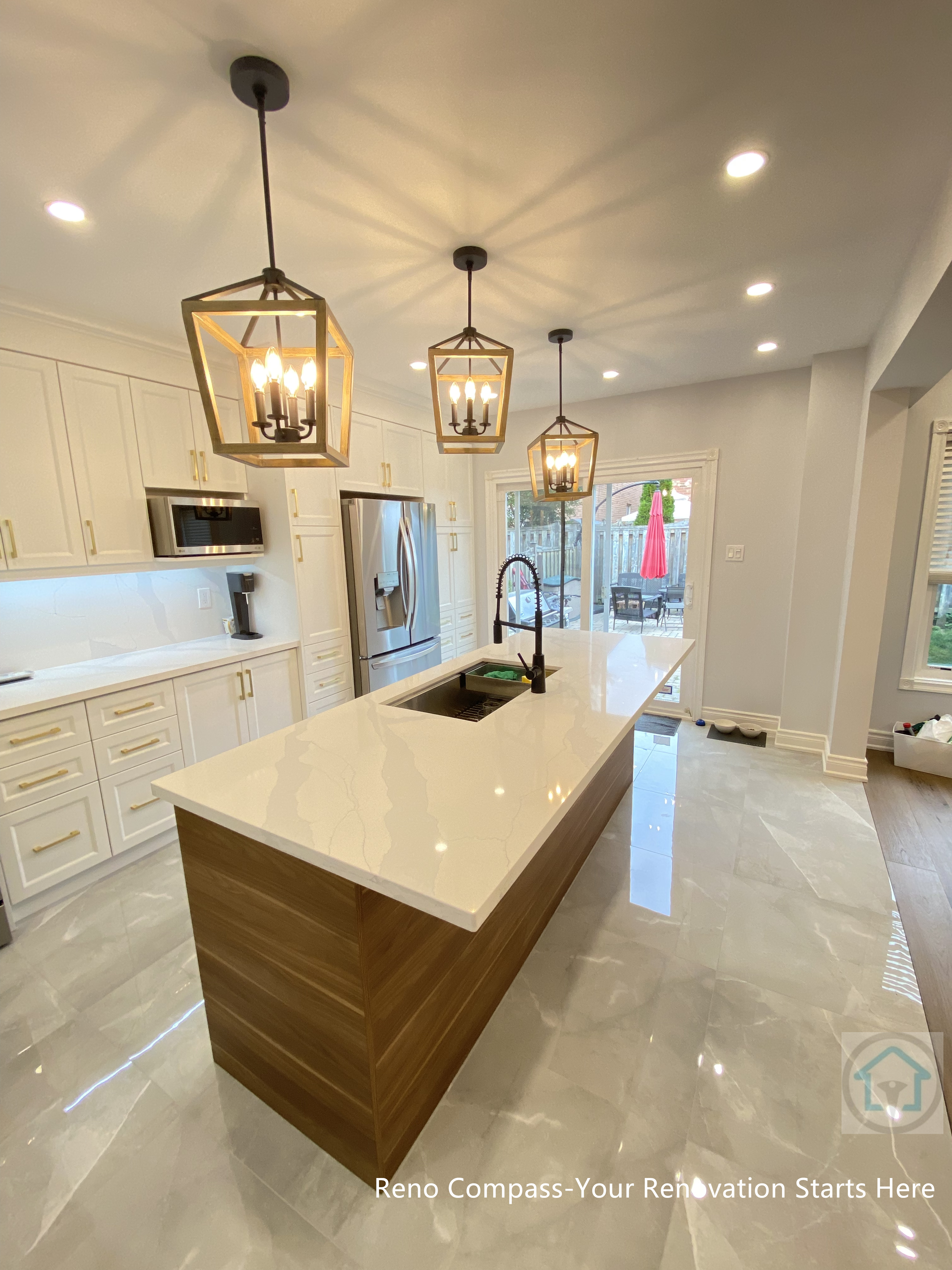 This image highlights a Newmarket interior renovation featuring a long quartz kitchen island with warm wood base cabinetry, designed to anchor the open-concept layout. The linear arrangement of lantern-style pendant lights creates strong visual rhythm and enhances the sense of depth within the space. White shaker cabinets, integrated stainless steel appliances, and glossy marble-look flooring work together to reflect light and elevate the overall brightness. A high-arc faucet and built-in sink add both functionality and modern appeal. Completed by a Toronto renovation company, this kitchen renovation emphasizes clean lines, balanced proportions, and seamless flow between cooking, dining, and outdoor access.