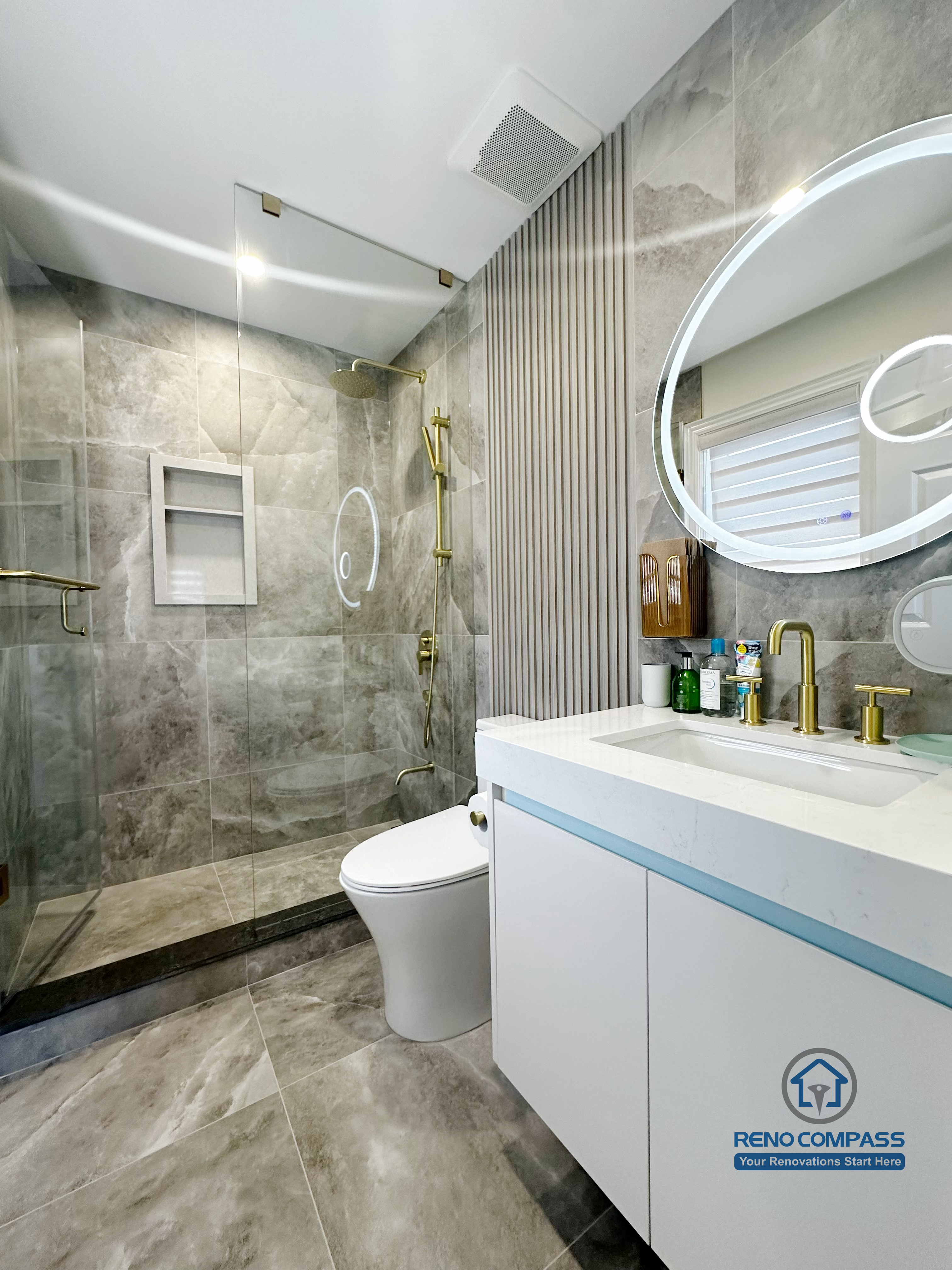 This image showcases a modern bathroom renovation in North York, featuring a full-height glass shower enclosure with large-format grey stone tiles. Brushed gold shower fixtures and built-in wall niches add both function and visual contrast, while the floating vanity with integrated sink enhances the clean, contemporary look. The combination of neutral stone surfaces, reflective glass, and warm metal accents creates a refined and cohesive bathroom design focused on durability and everyday comfort.