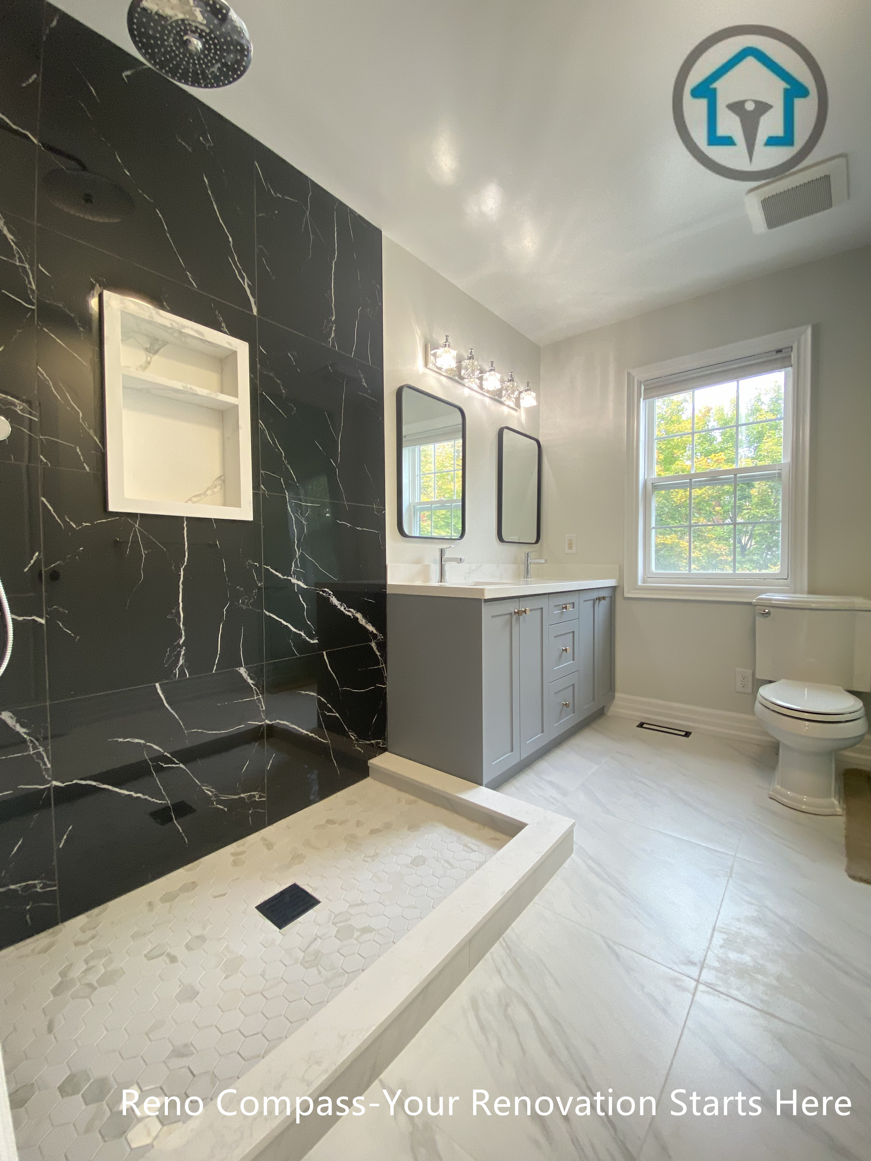This modern bathroom renovation features a sleek curbless walk-in shower with black marble-look wall tiles, creating a bold contrast against the light flooring. A custom double vanity with integrated storage and clean-lined mirrors enhances daily functionality while maintaining a refined, contemporary aesthetic. Natural light from the window balances the dark accent wall, resulting in a bathroom design that feels both elegant and practical.