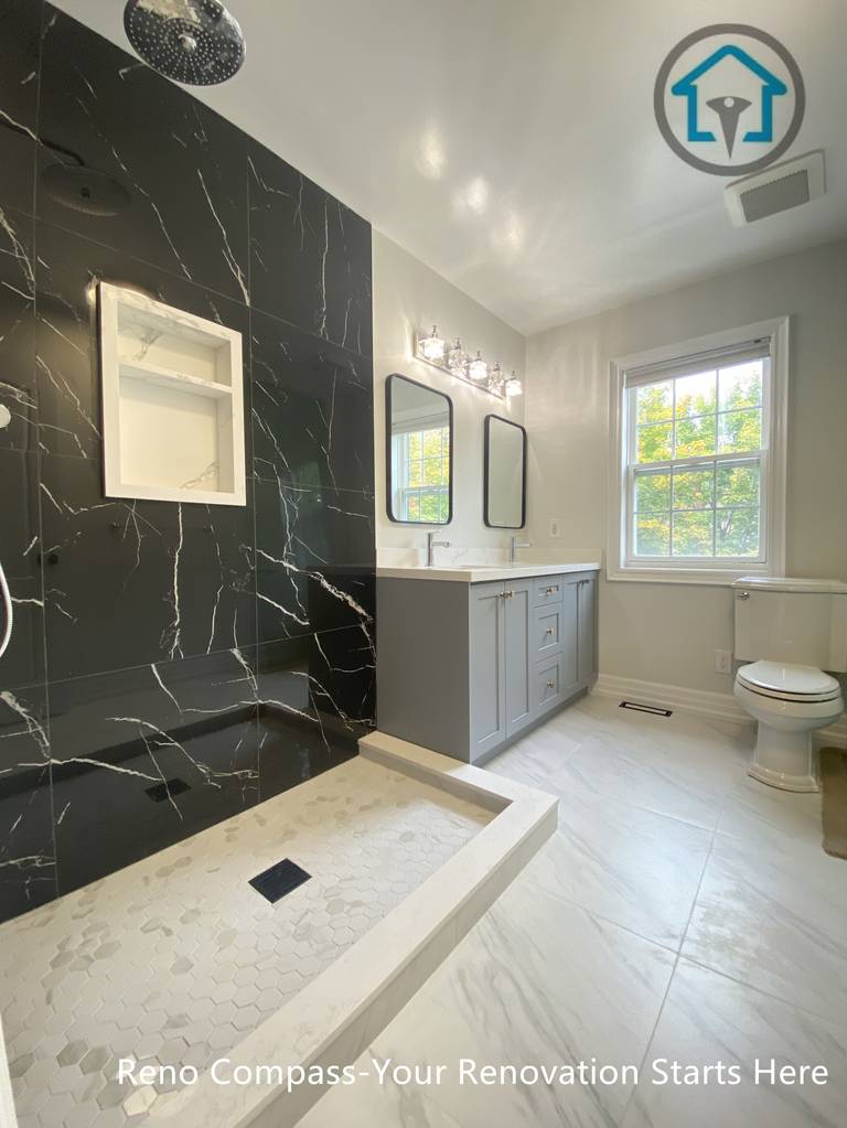 This modern bathroom renovation features a sleek curbless walk-in shower with black marble-look wall tiles, creating a bold contrast against the light flooring. A custom double vanity with integrated storage and clean-lined mirrors enhances daily functionality while maintaining a refined, contemporary aesthetic. Natural light from the window balances the dark accent wall, resulting in a bathroom design that feels both elegant and practical.