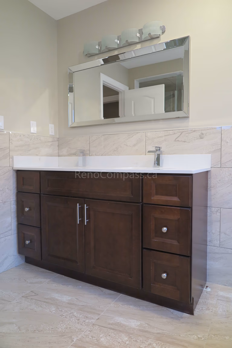 This Markham bathroom renovation features a custom dark wood vanity with a sleek quartz countertop and integrated sink, creating a clean and functional layout. Marble-style wall and floor tiles enhance the elegant and modern design, while updated lighting and mirror installation complete the refined look. Completed by Reno Compass, a professional renovation contractor specializing in bathroom renovations, vanity installation, and full interior remodeling across Markham and the Greater Toronto Area.