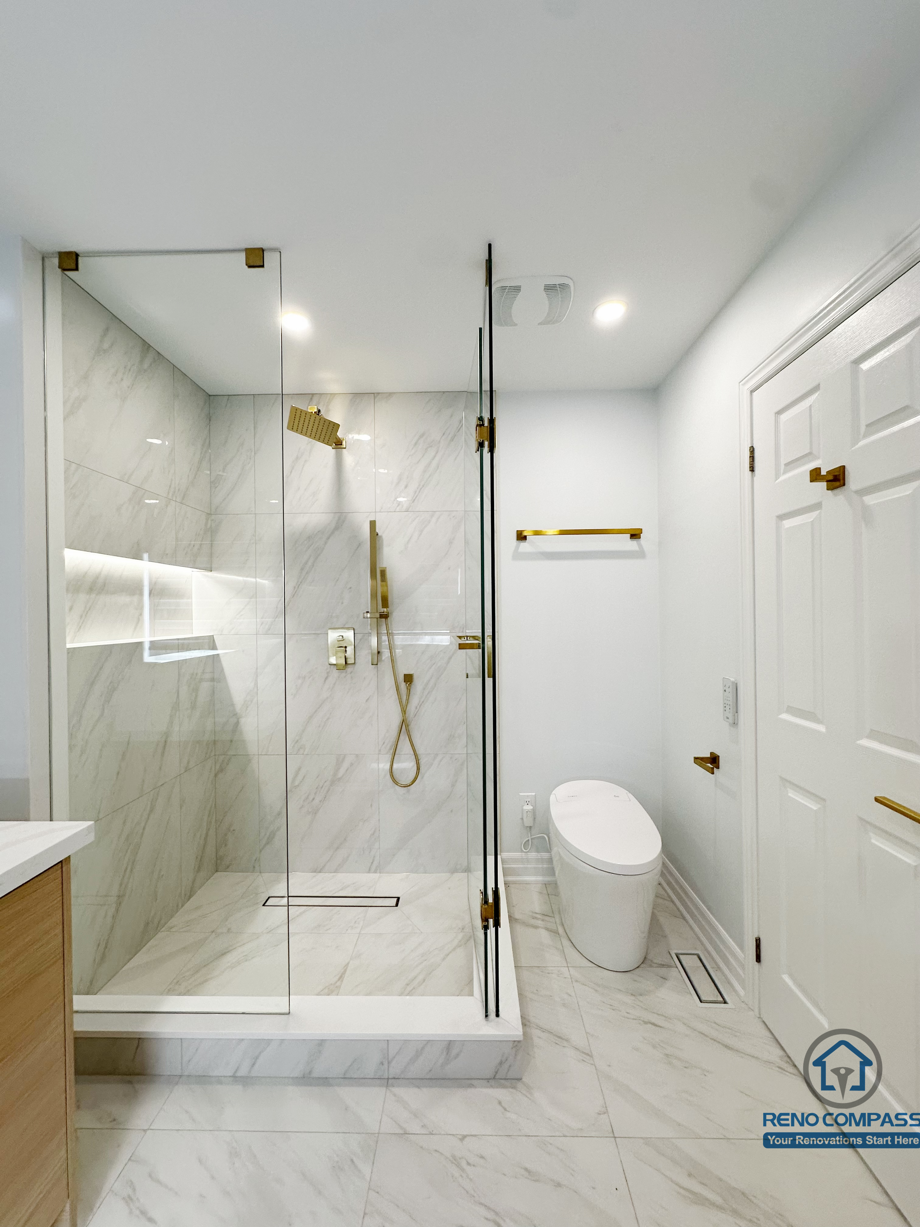 Contemporary bathroom renovation featuring a frameless glass walk-in shower, marble-look wall and floor tiles, built-in shower niche, and elegant gold-tone fixtures for a clean, modern finish.